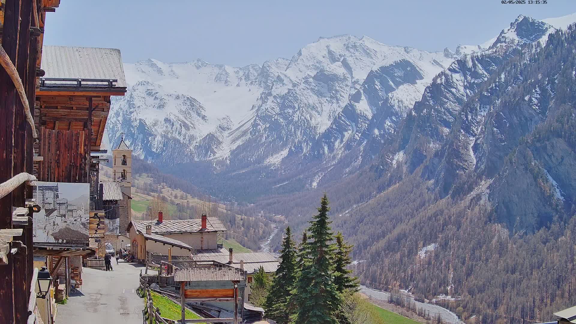 A sunny view of a small village nestled in a valley, overlooked by snow-capped mountains.