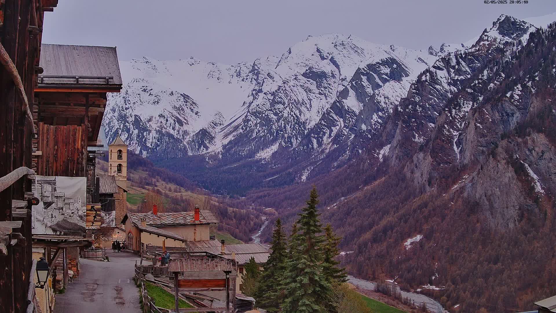 A mountain village with wooden buildings and a church overlooks a valley and snow-capped mountains under an overcast sky.