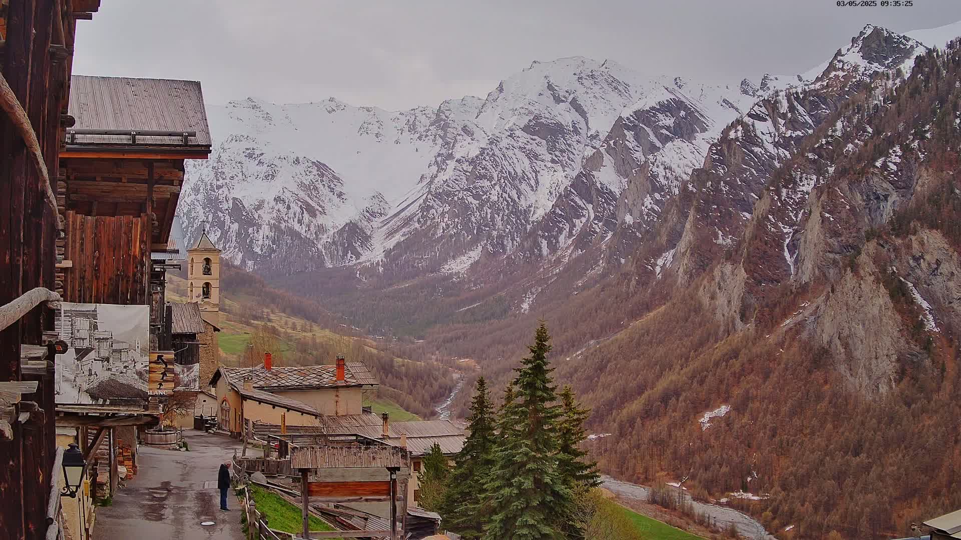 A person stands on a street in a mountain village overlooking a valley with a river and snow-capped mountains under an overcast sky.