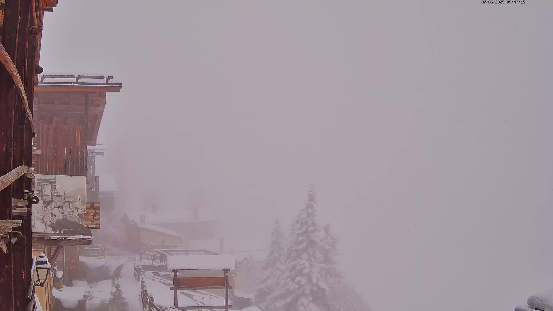 Snow falls heavily over a snow-covered village of wooden buildings, obscuring the view in a dense fog.