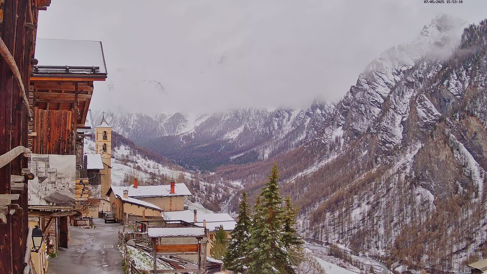 A snow-covered village nestled in a valley between towering, snow-dusted mountains under an overcast sky.