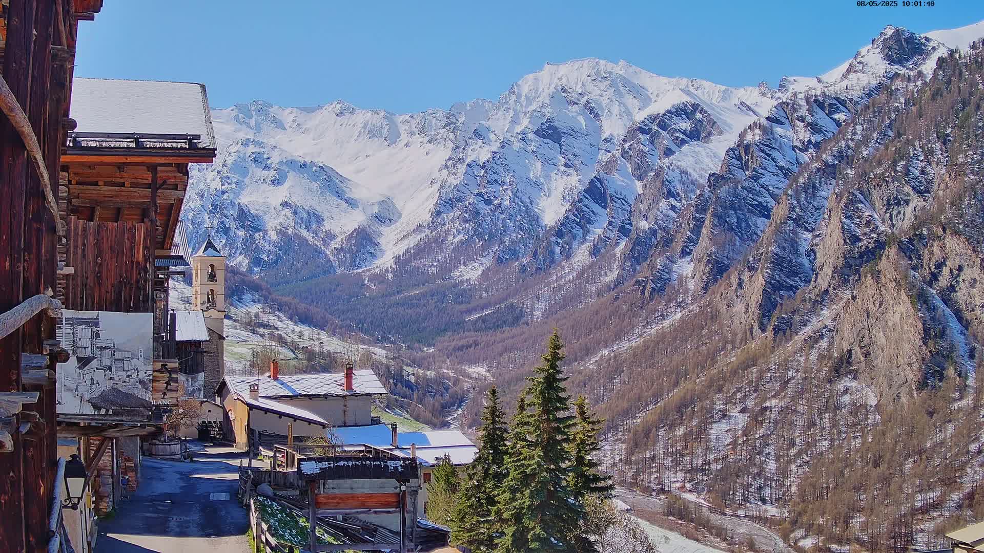A small village with snow-covered roofs nestles in a valley, overlooked by towering snow-capped mountains under a clear blue sky.