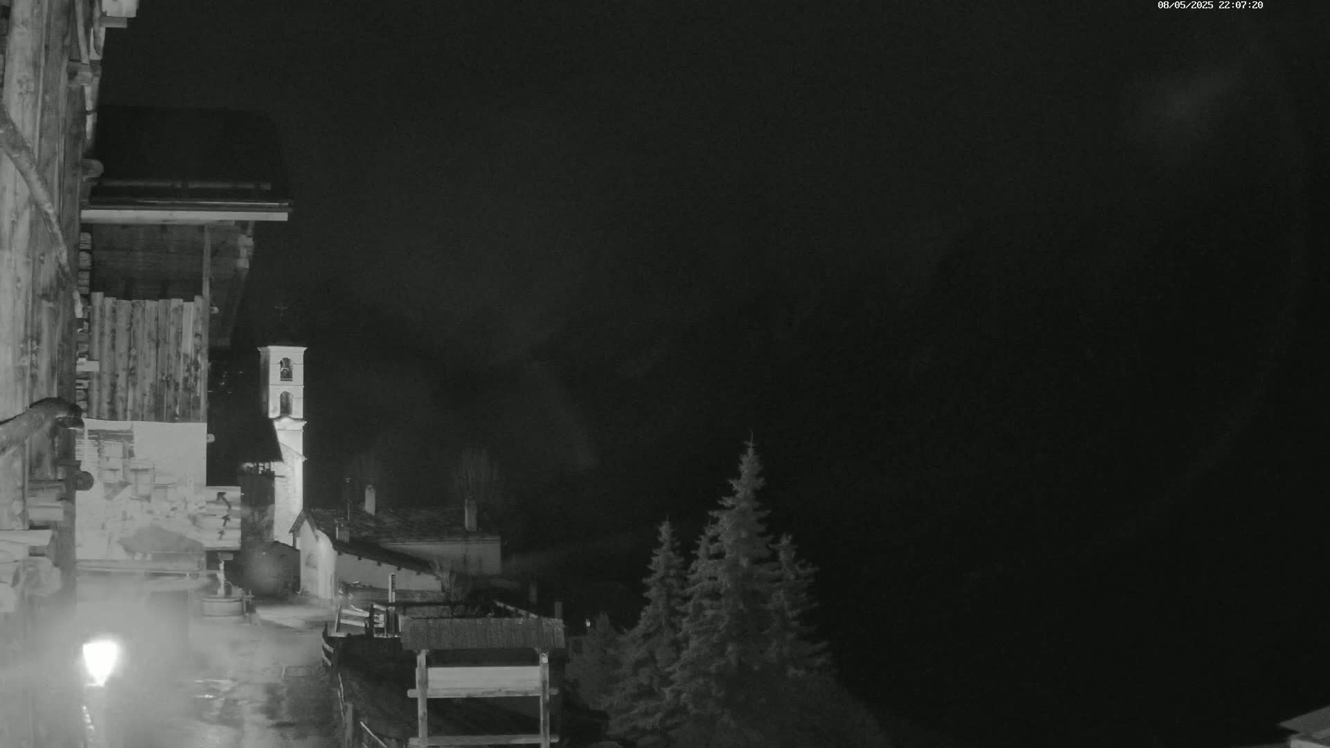 A nighttime view of a village with a bell tower, illuminated by a streetlight, under a dark, possibly cloudy sky.