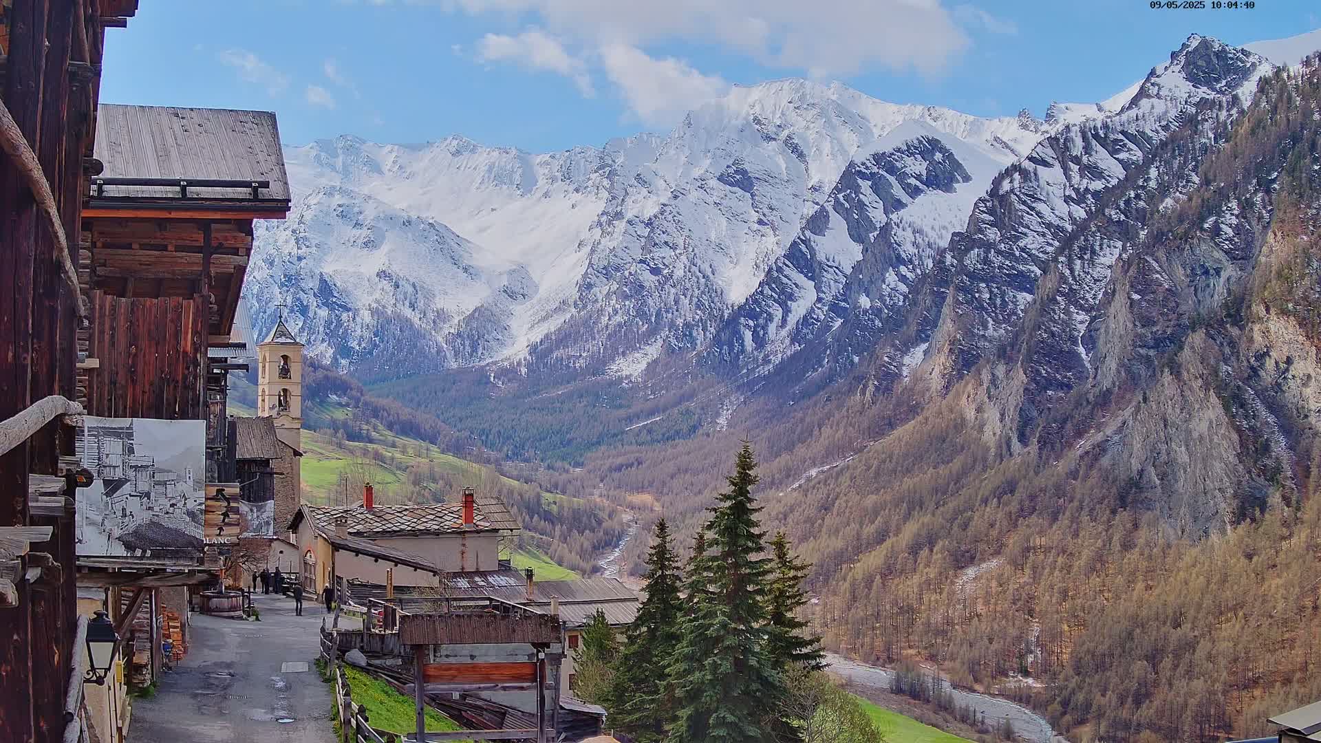 A quaint village nestled in a valley, overlooked by snow-capped mountains under a partly cloudy sky.