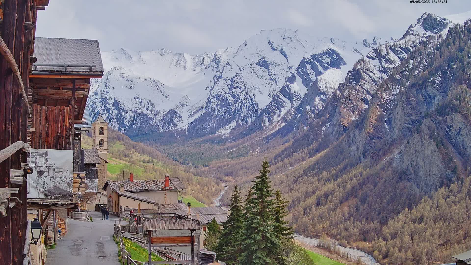 A mountain village nestled in a valley, with snow-capped mountains in the background under a partly cloudy sky.