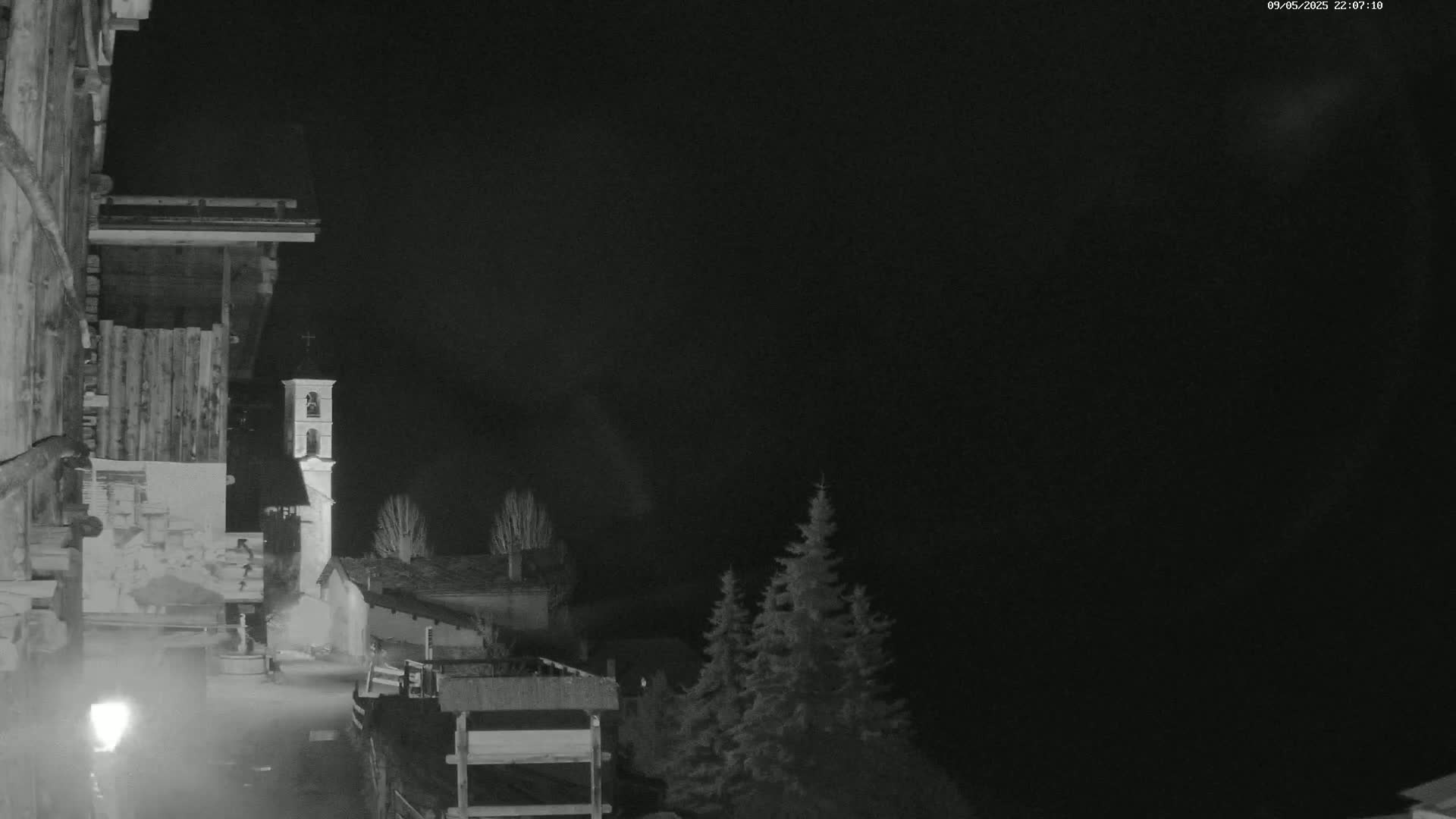A nighttime view of a village scene, featuring a church bell tower, buildings, and evergreens under a dark, cloudy sky.