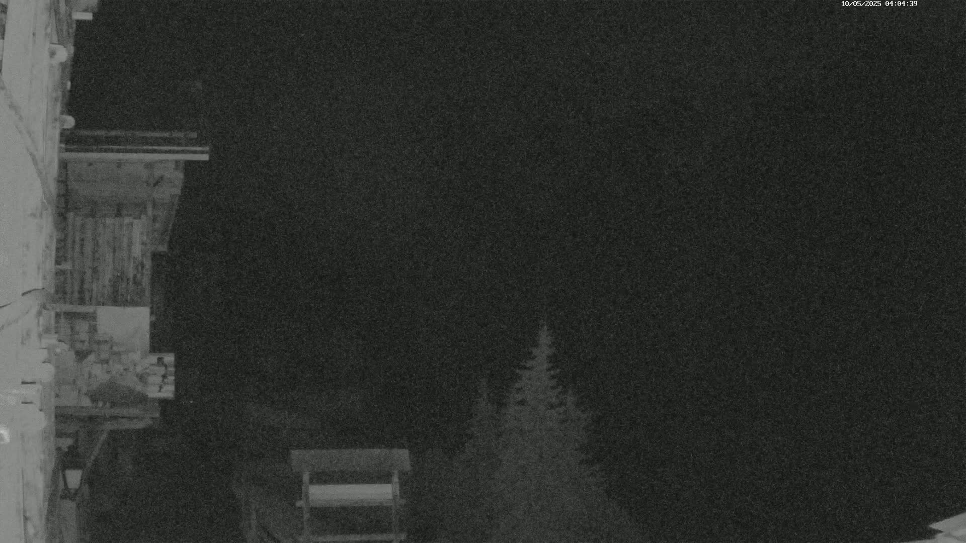 A nighttime, low-light image shows a wooden balcony attached to a building, with a few dark evergreen trees visible in the background under a dark, clear sky.