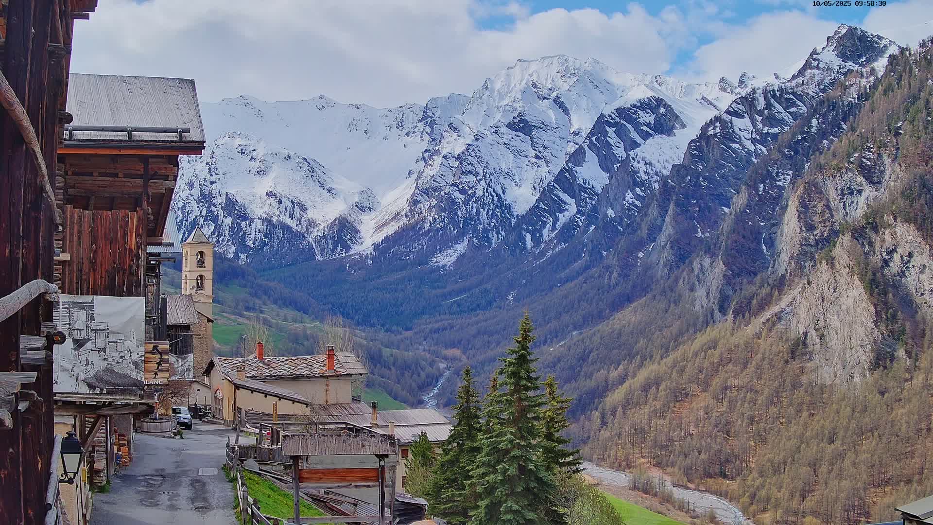 A quaint mountain village with wooden buildings nestles in a valley, overlooked by snow-capped mountains under a partly cloudy sky.