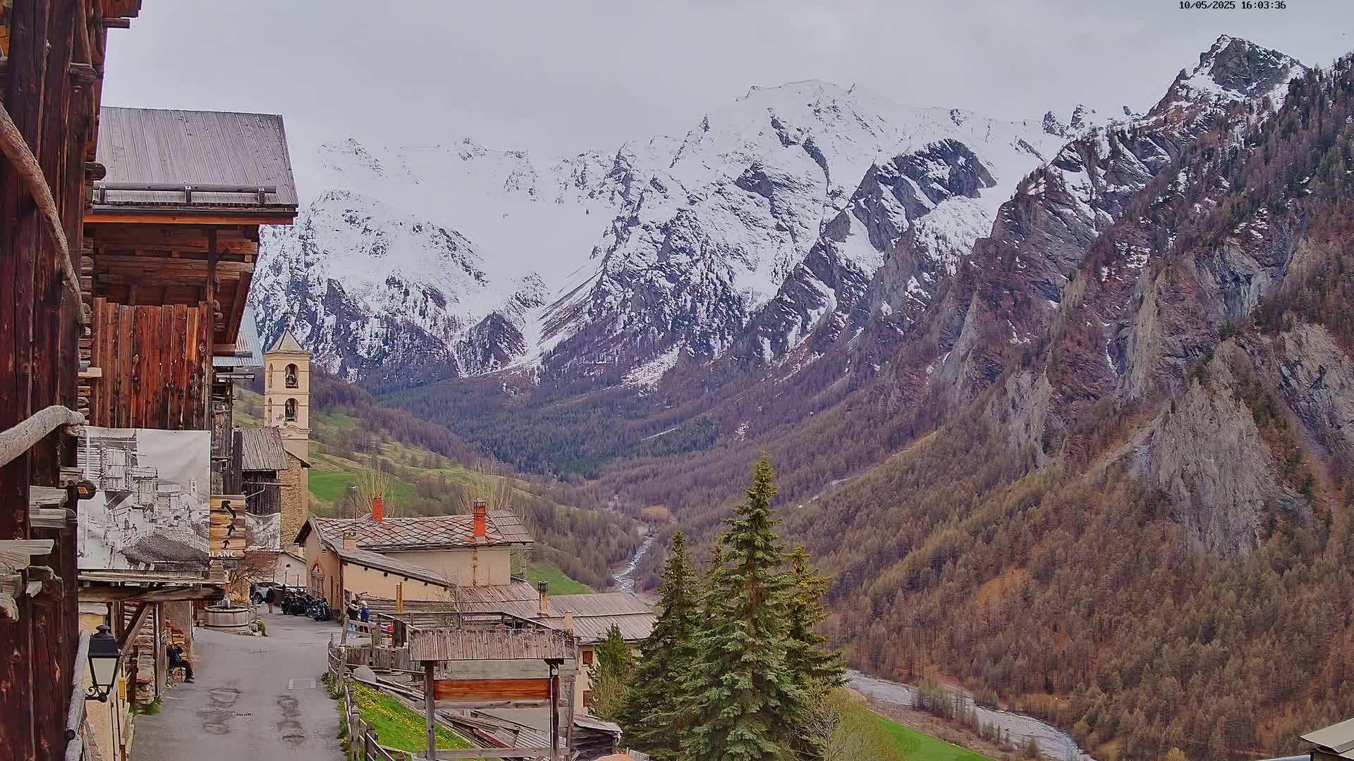 A mountain village with aged wooden buildings sits nestled in a valley, overlooking a snow-capped mountain range under an overcast sky.