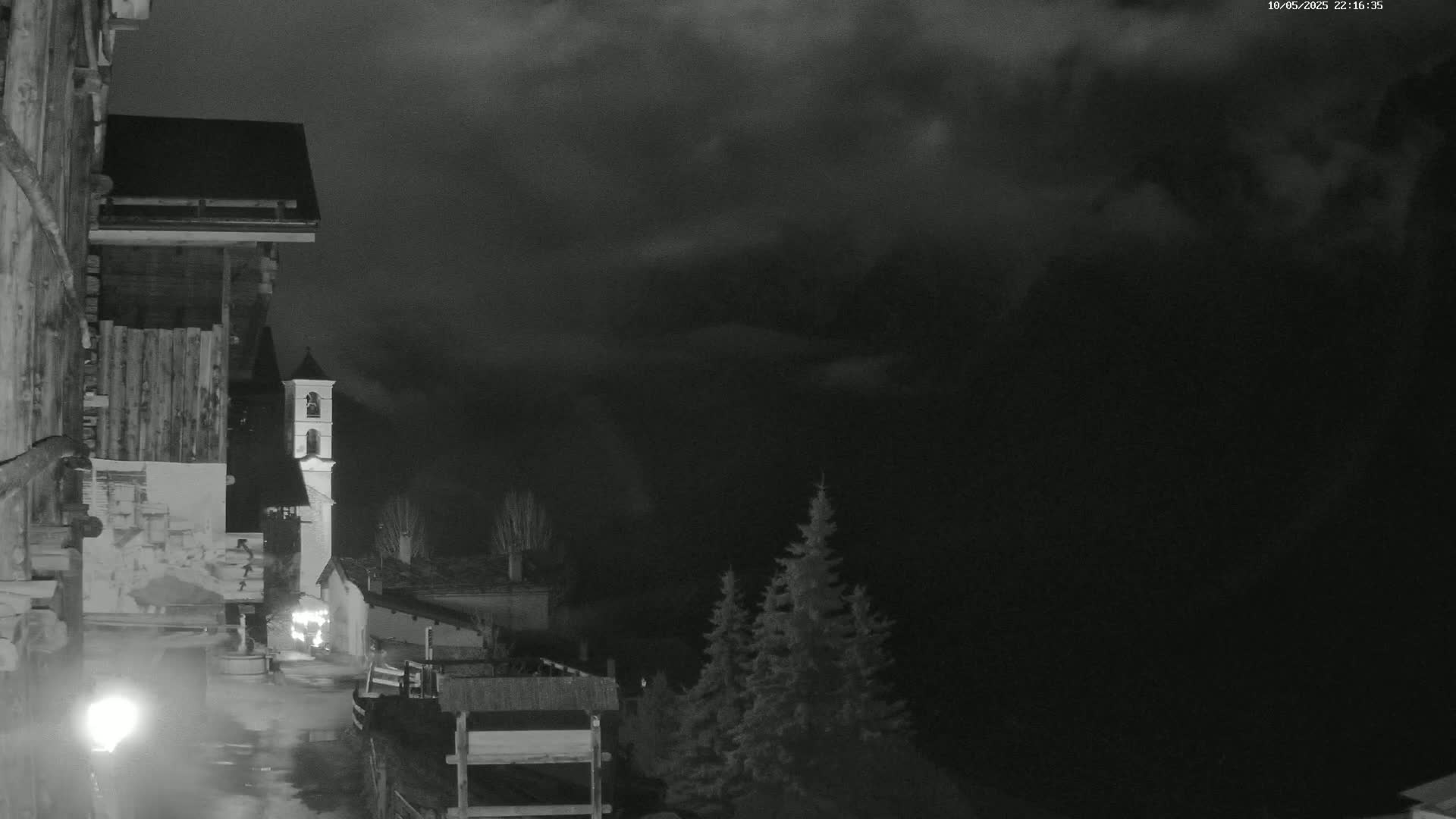A nighttime view of a village nestled in a valley under a cloudy sky, showing a church steeple and traditional buildings.