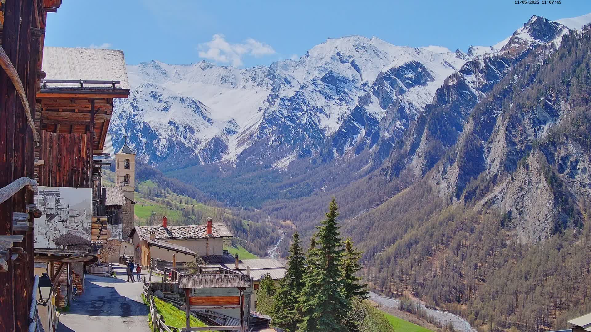 A mountain village nestled in a valley, with snow-capped mountains in the background under a clear blue sky.