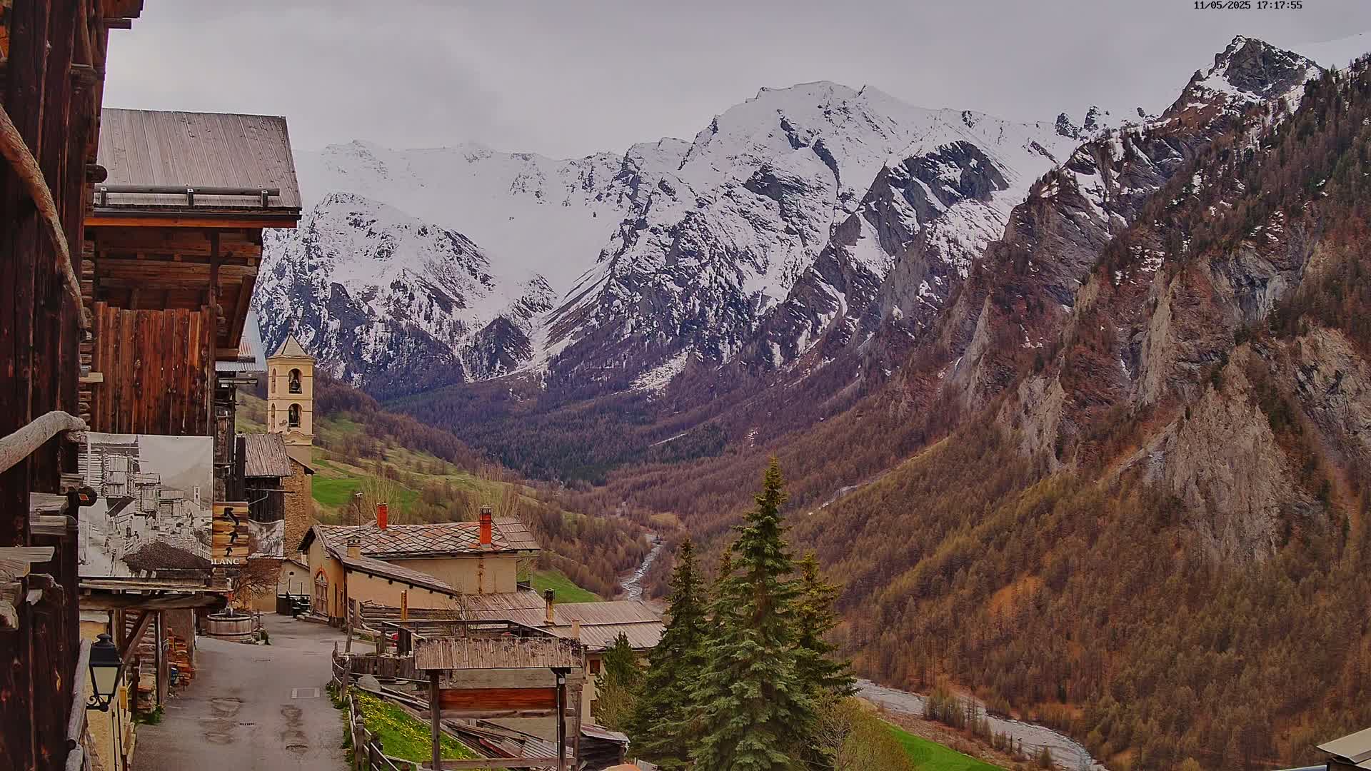 A snow-capped mountain range overlooks a small village of rustic stone buildings nestled in a valley under an overcast sky.