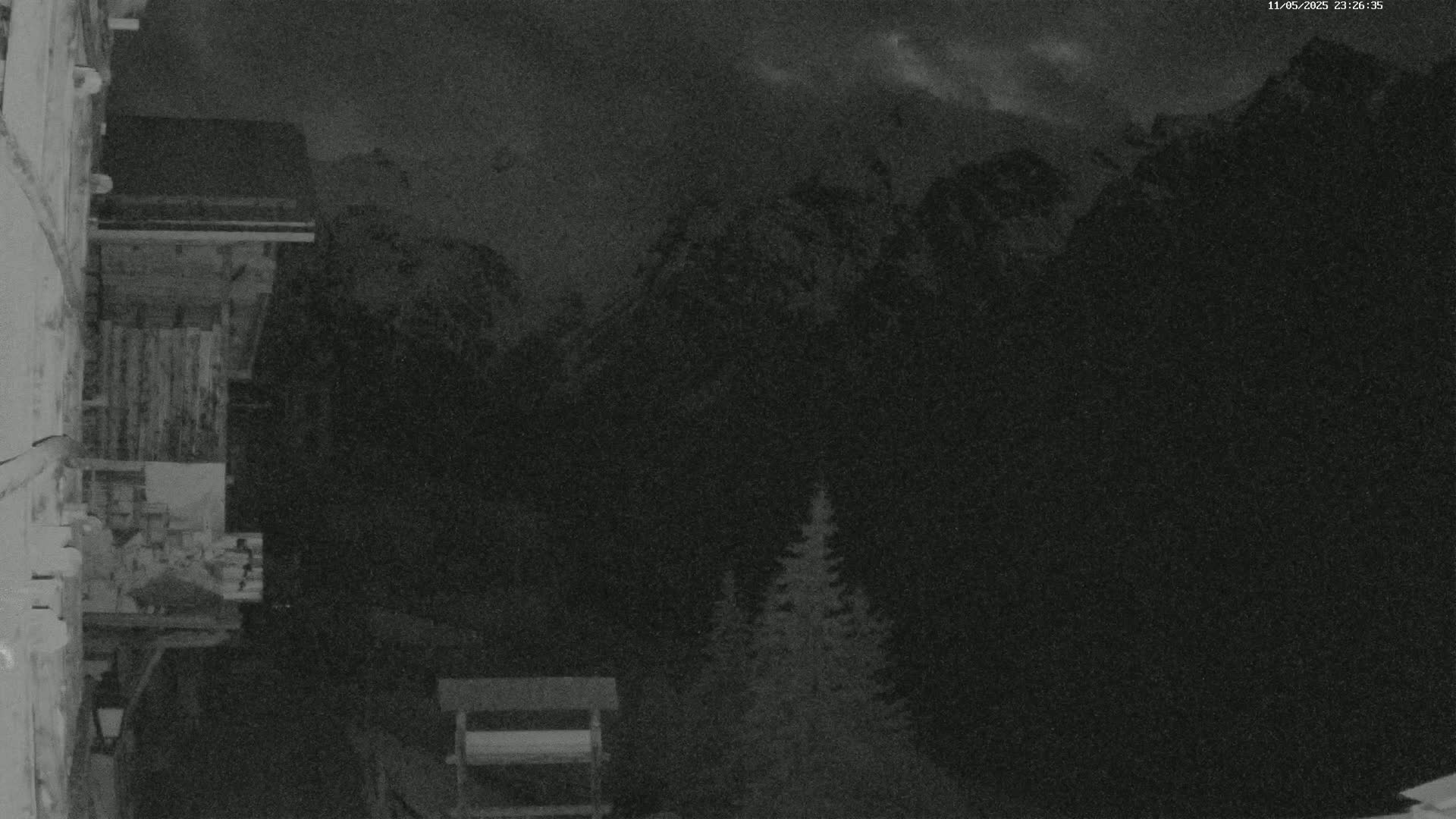 A nighttime, low-light image shows a wooden structure attached to a building overlooking a dark mountain range under a cloudy sky, with a small evergreen tree visible in the foreground.