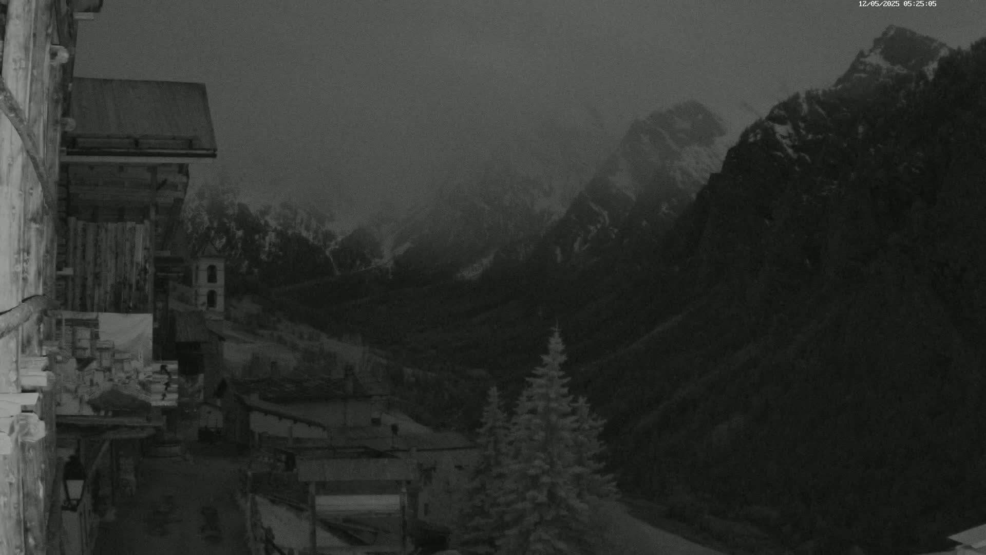 A dark, overcast view from a mountain village shows snow-dusted peaks in the distance.
