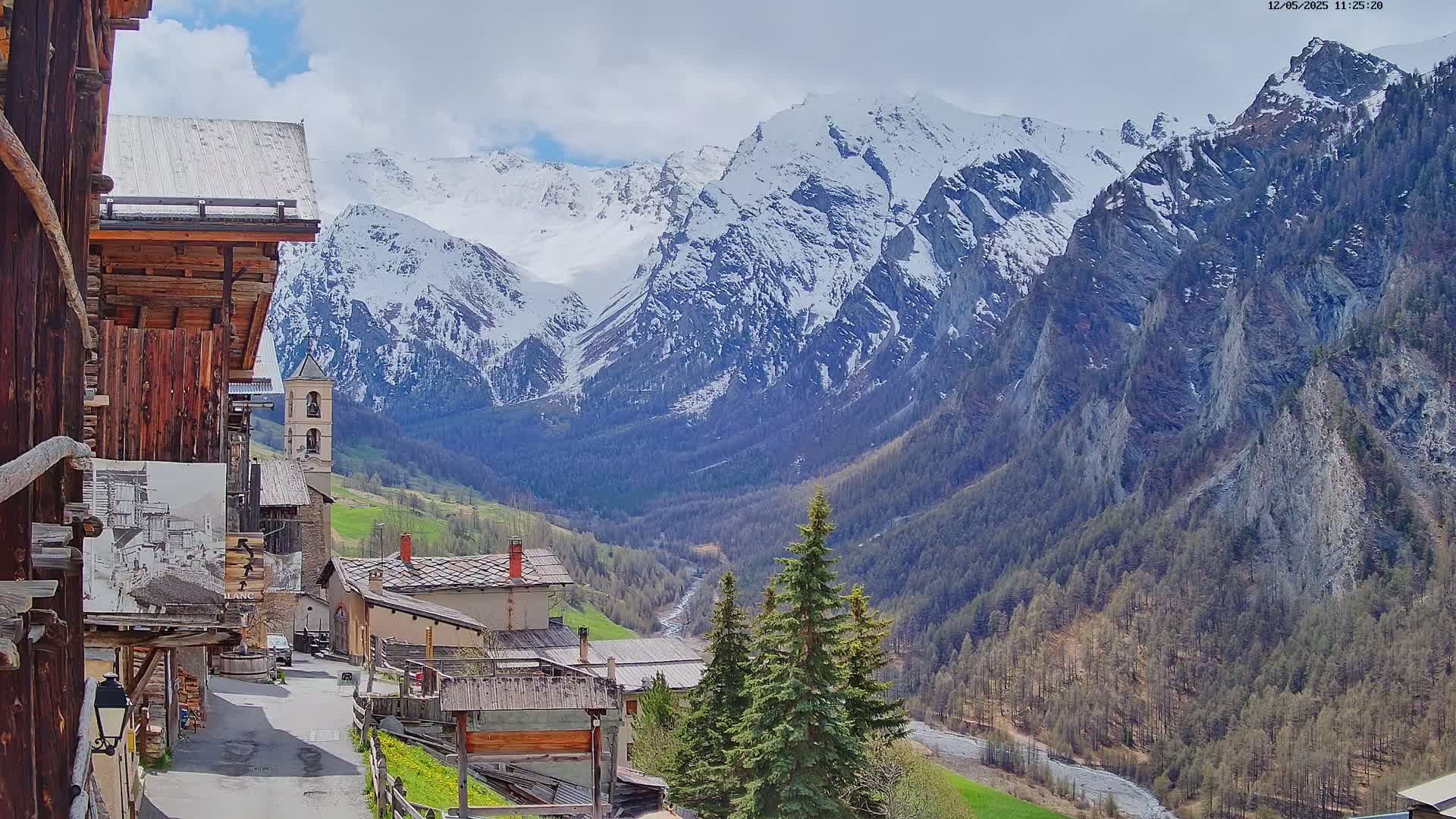 A quaint mountain village nestled in a valley, with snow-capped mountains rising in the background under a partly cloudy sky.