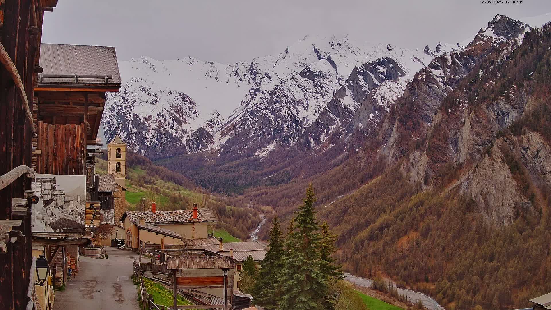 A village of stone and wood buildings nestled in a valley, overlooked by snow-capped mountains under a cloudy sky.
