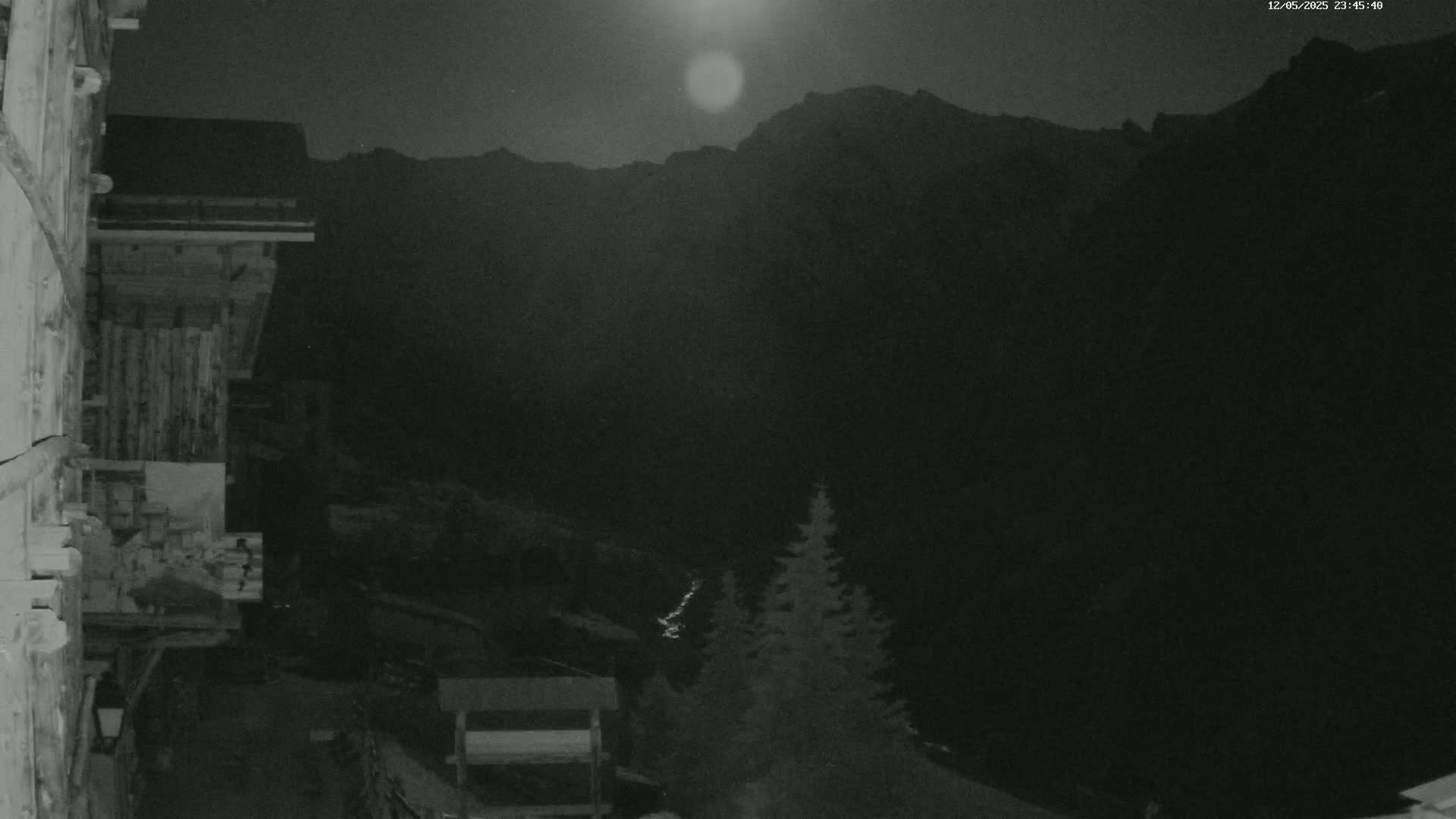 A nighttime view from a wooden building shows a moonlit mountainous landscape with a few evergreens in the foreground.