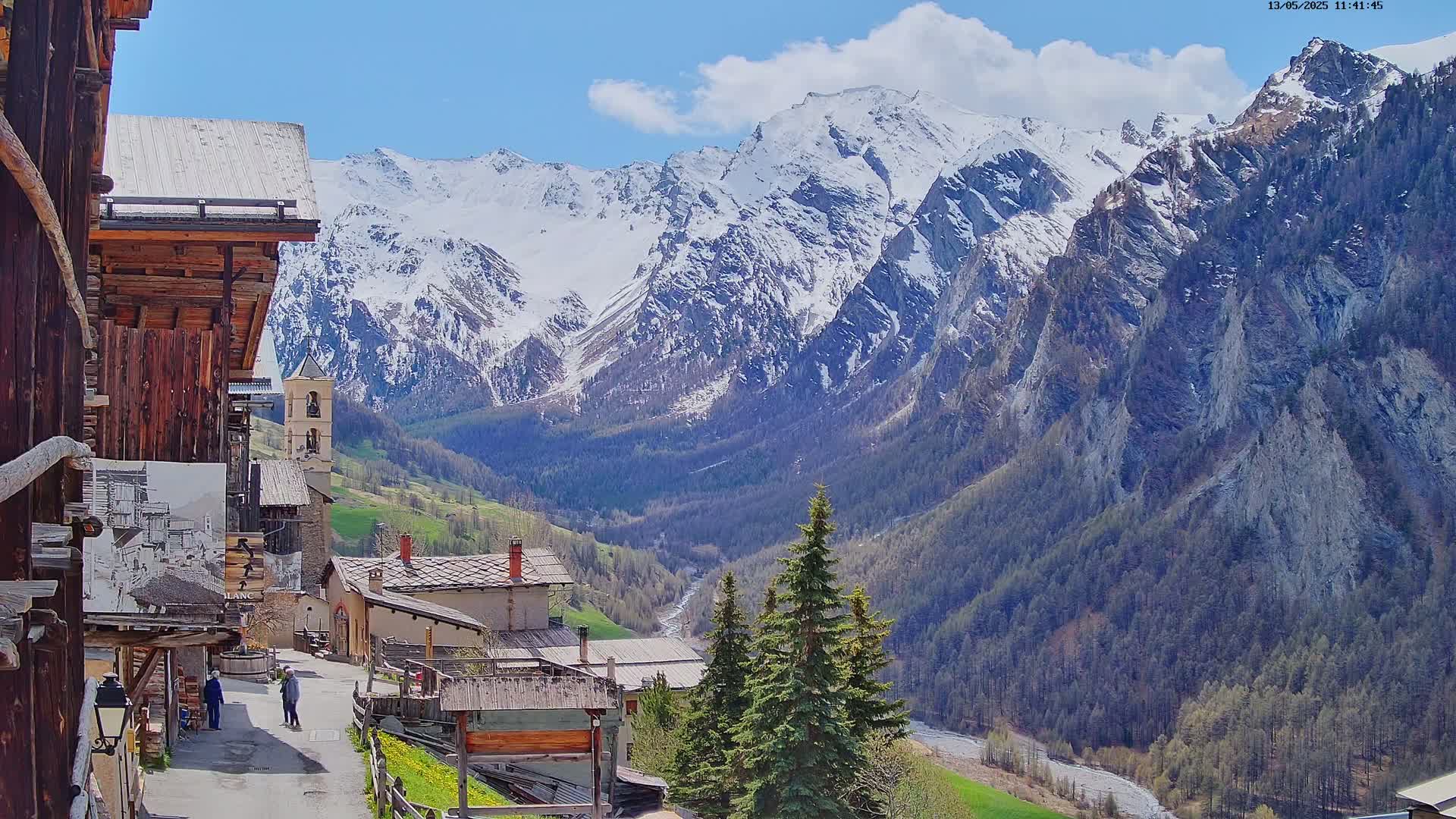 A quaint mountain village with stone and wood buildings is nestled in a valley, overlooked by snow-capped mountains under a mostly sunny sky.