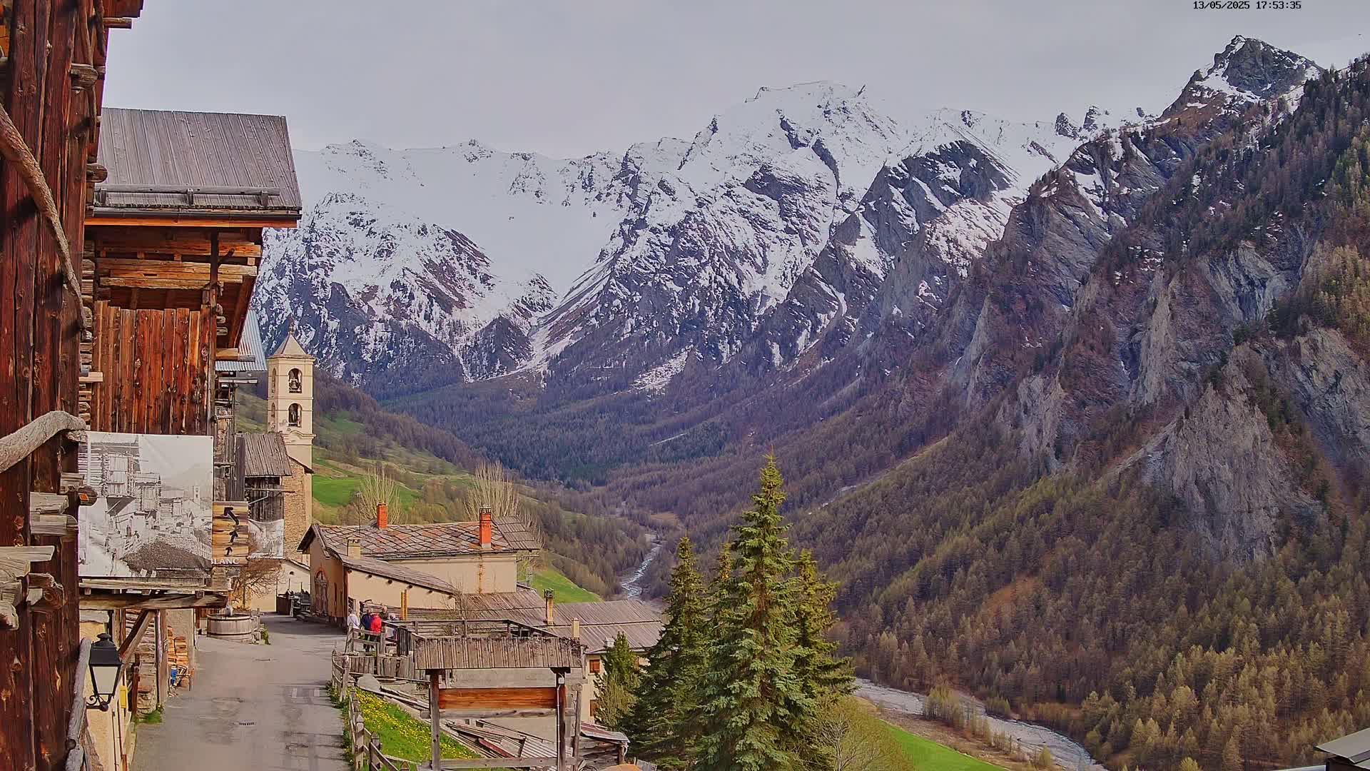 A mountain village with aged wooden buildings is nestled in a valley, overlooked by snow-capped mountains under a cloudy sky.