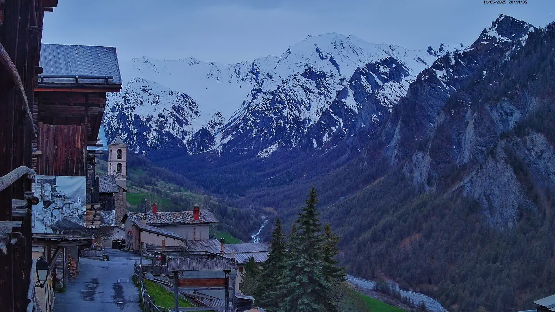 A mountain village with dark wooden buildings and a church overlooks a valley with a river flowing through it, all set against a backdrop of snow-capped mountains under a twilight sky.