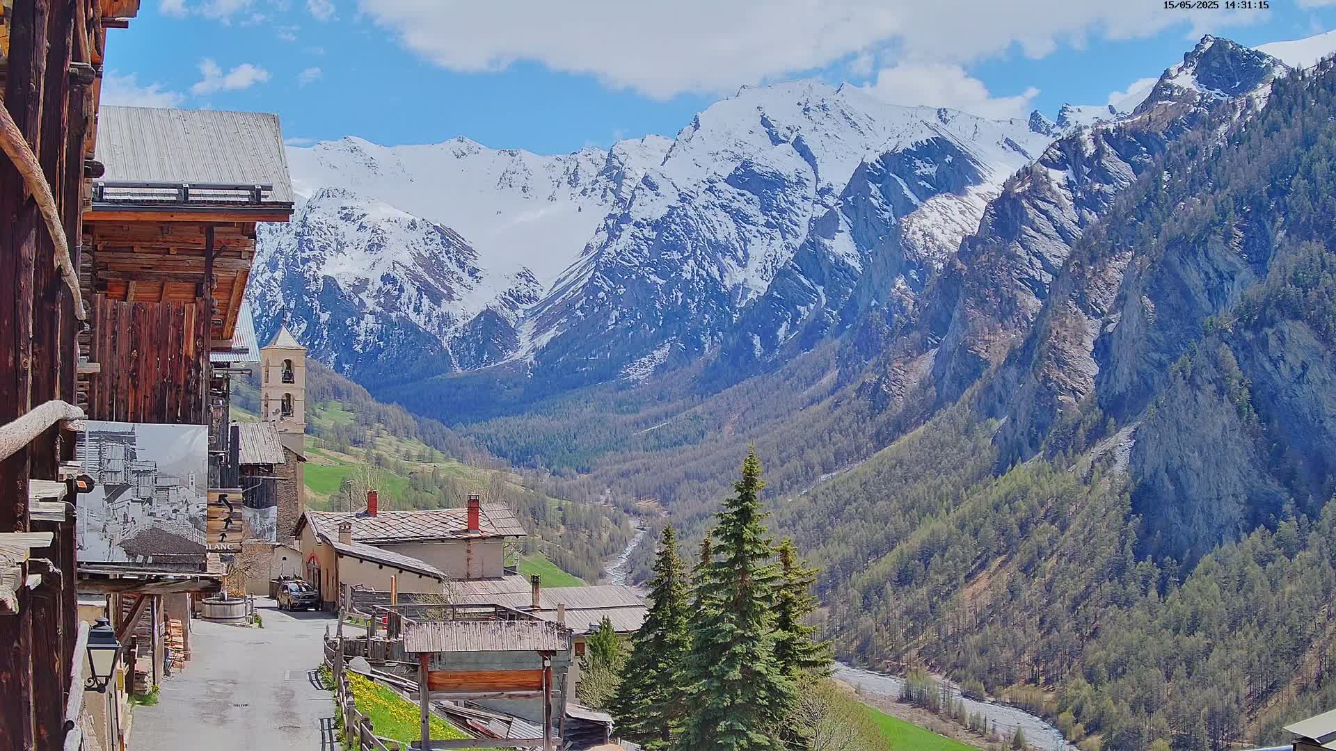 A quaint mountain village with wooden buildings nestles in a valley, overlooked by snow-capped mountains under a mostly sunny sky.