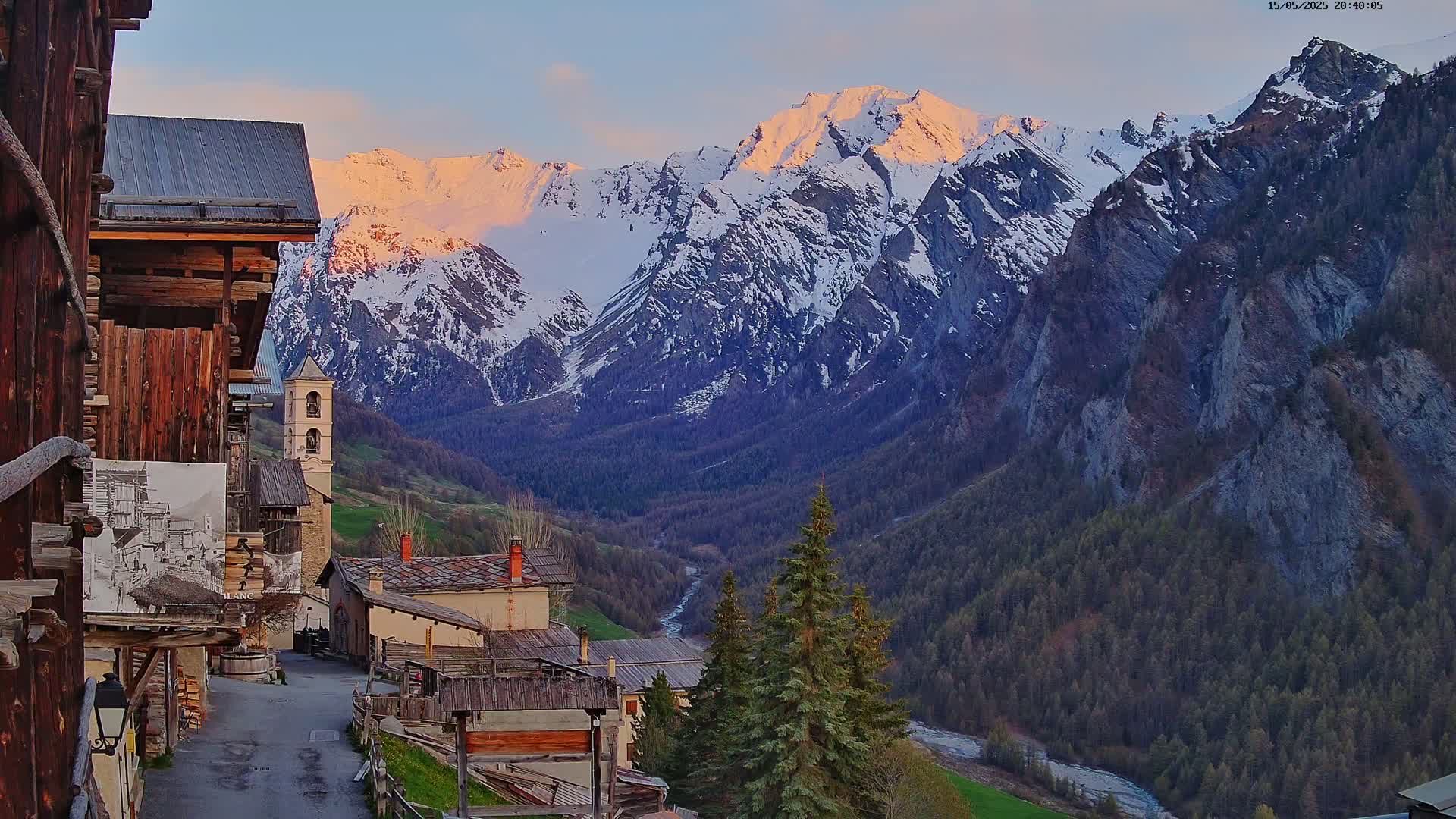 A quaint mountain village nestled in a valley, with a sunlit snow-capped mountain range in the background, at sunset.
