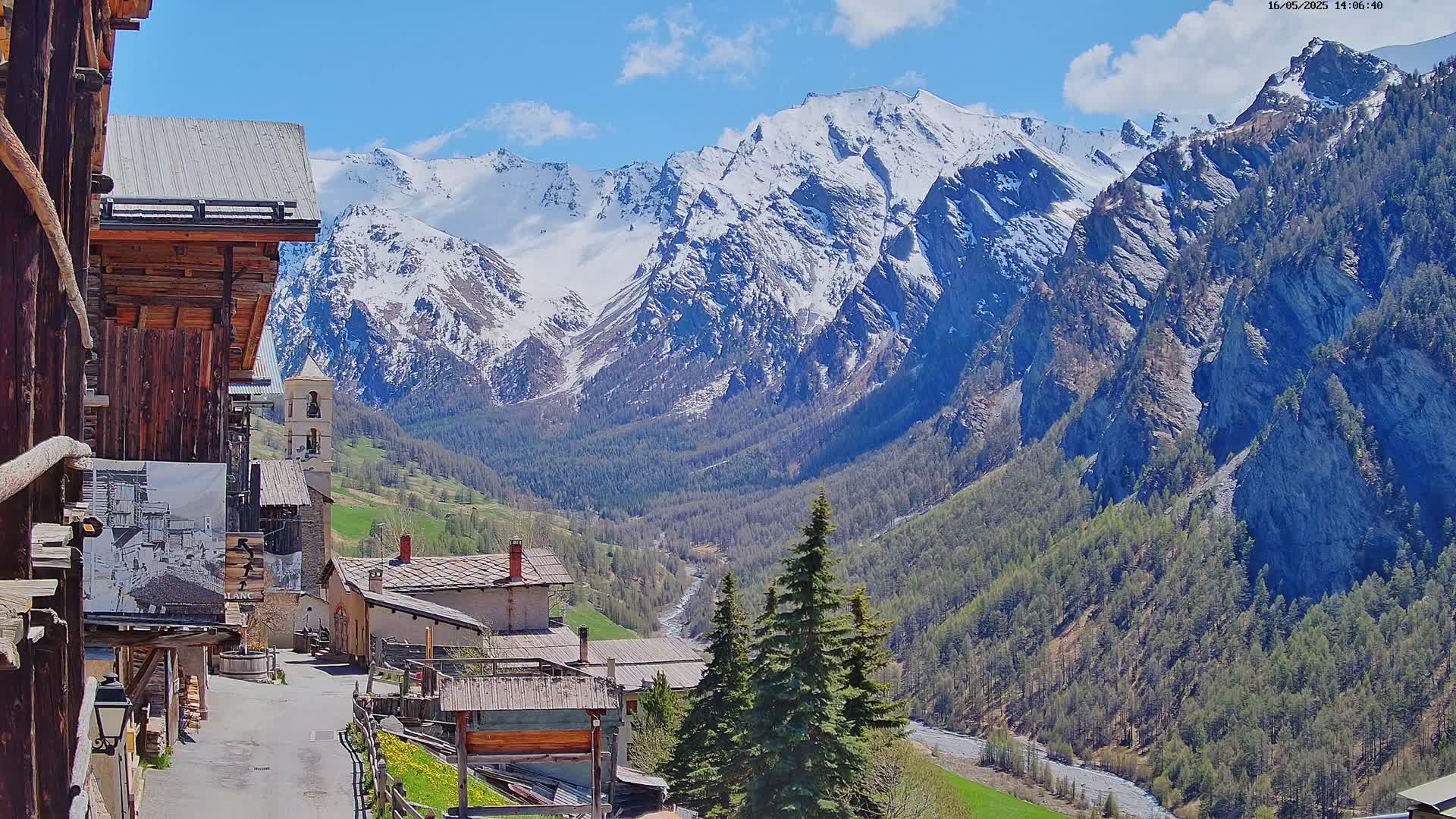 A sunny view from a wooden building shows a small village nestled in a valley, with snow-capped mountains rising in the background.