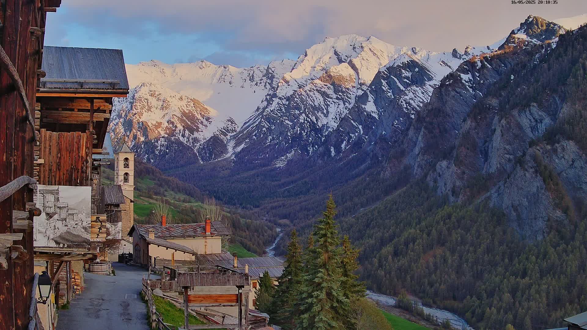 A quaint mountain village nestled in a valley, overlooked by snow-capped mountains under a partly cloudy evening sky.