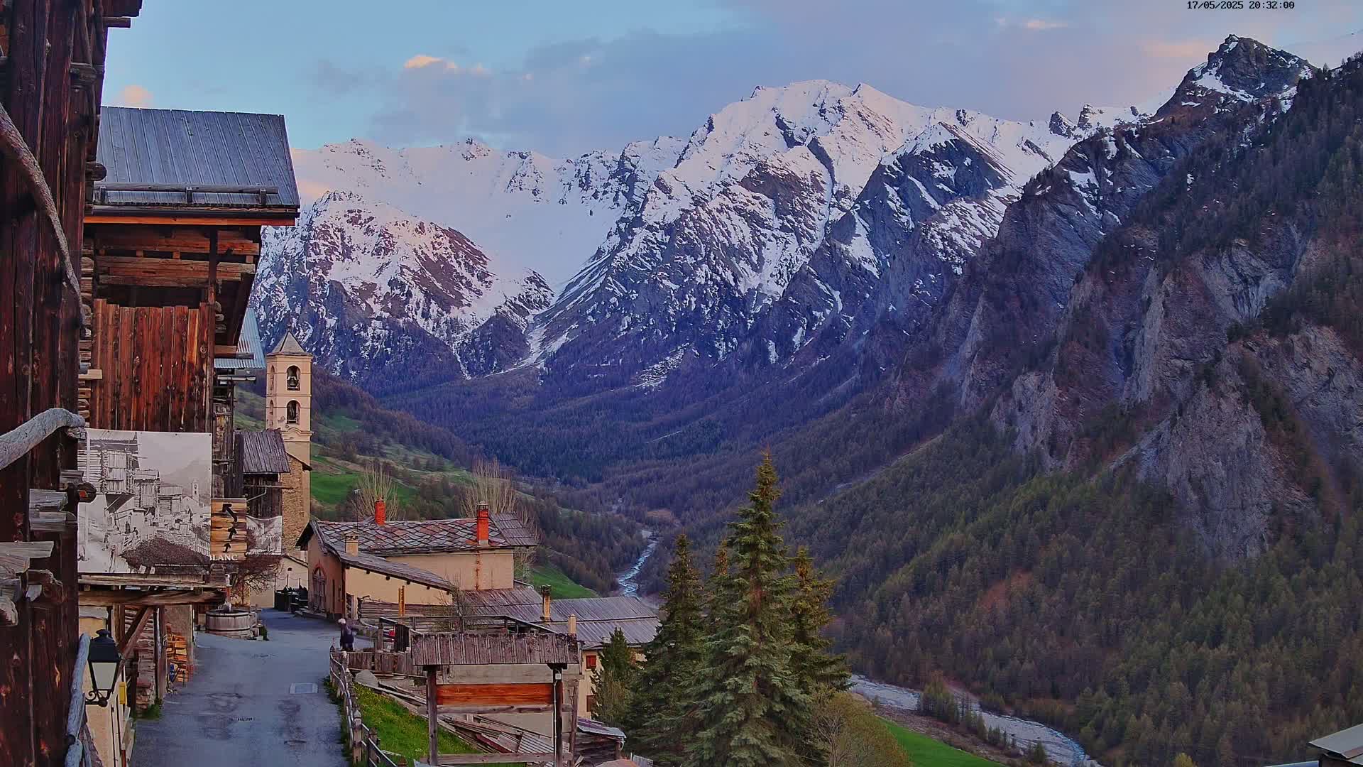 A quaint mountain village nestled in a valley, overlooked by snow-capped mountains under a twilight sky.