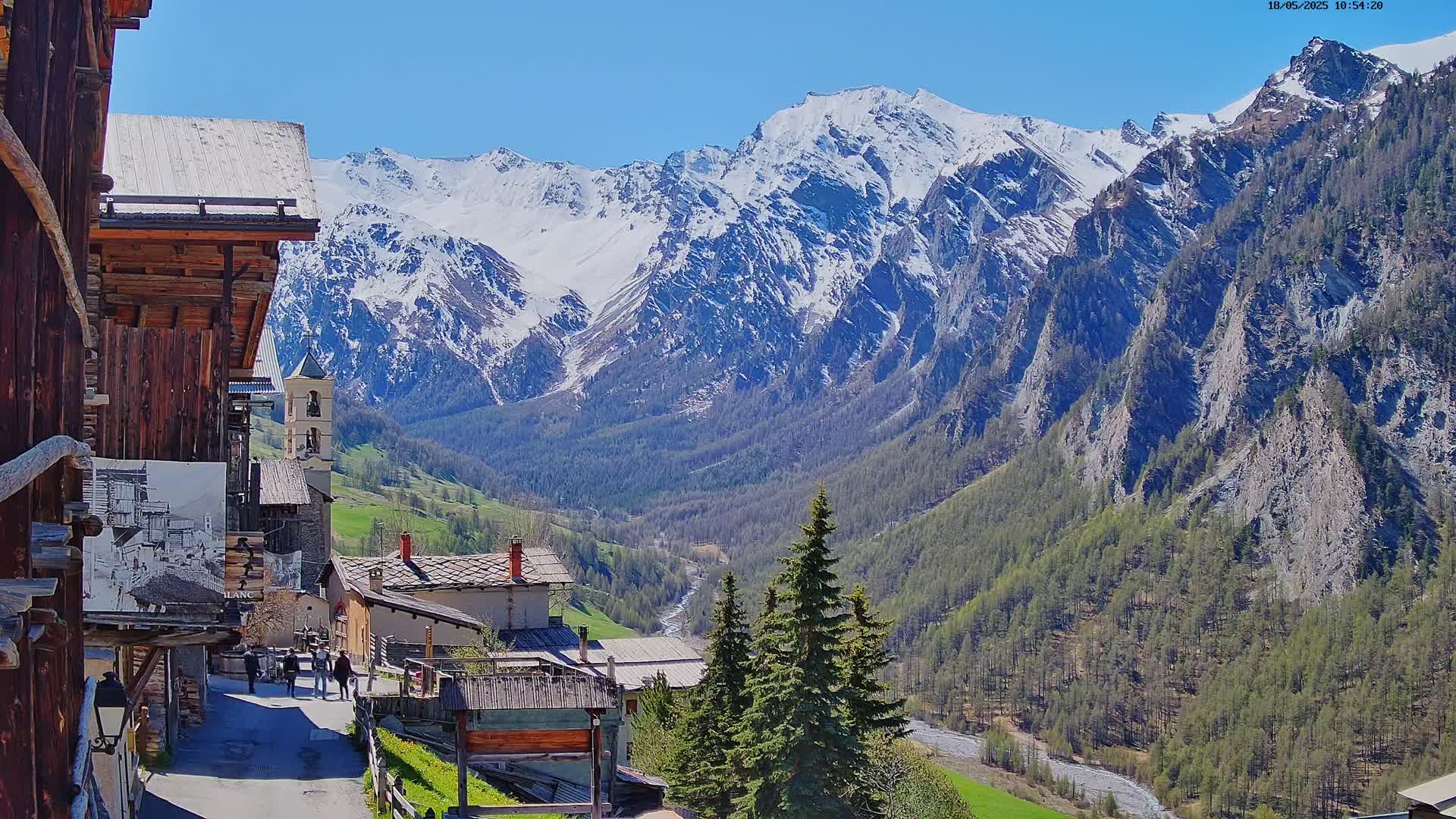A sunny mountain village nestled in a valley, surrounded by snow-capped peaks and evergreen forests.
