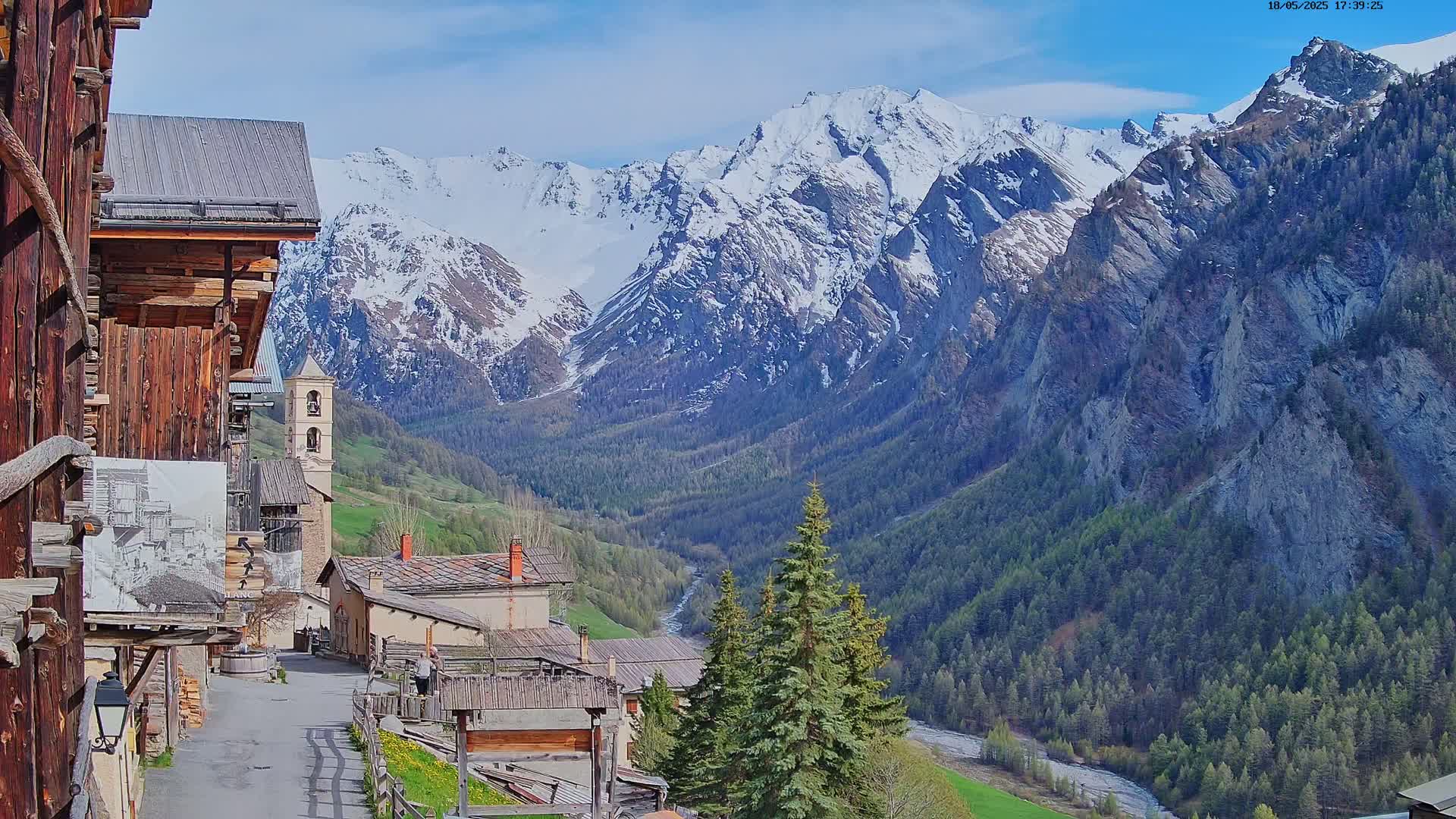 A quaint village nestled in a valley, surrounded by snow-capped mountains under a mostly sunny sky.