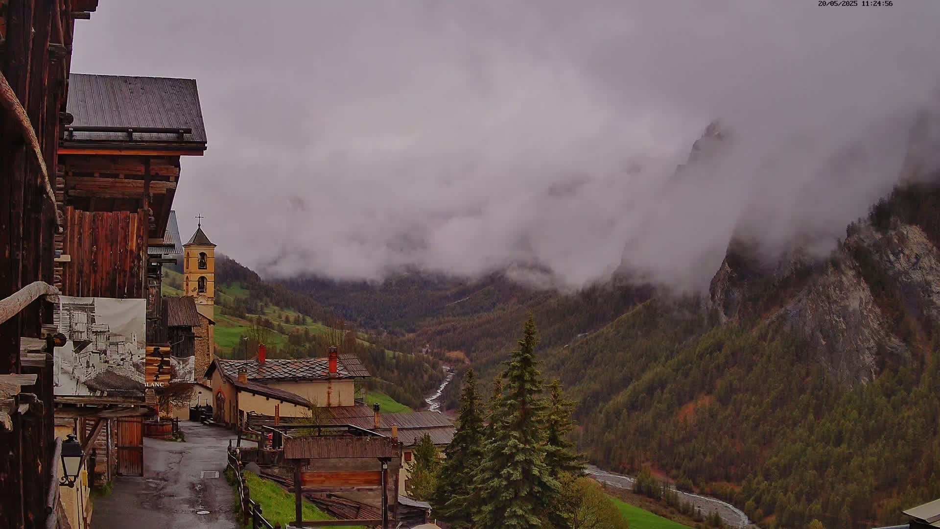 A misty mountain valley view from a village with old wooden buildings, a church steeple, and a flowing river.