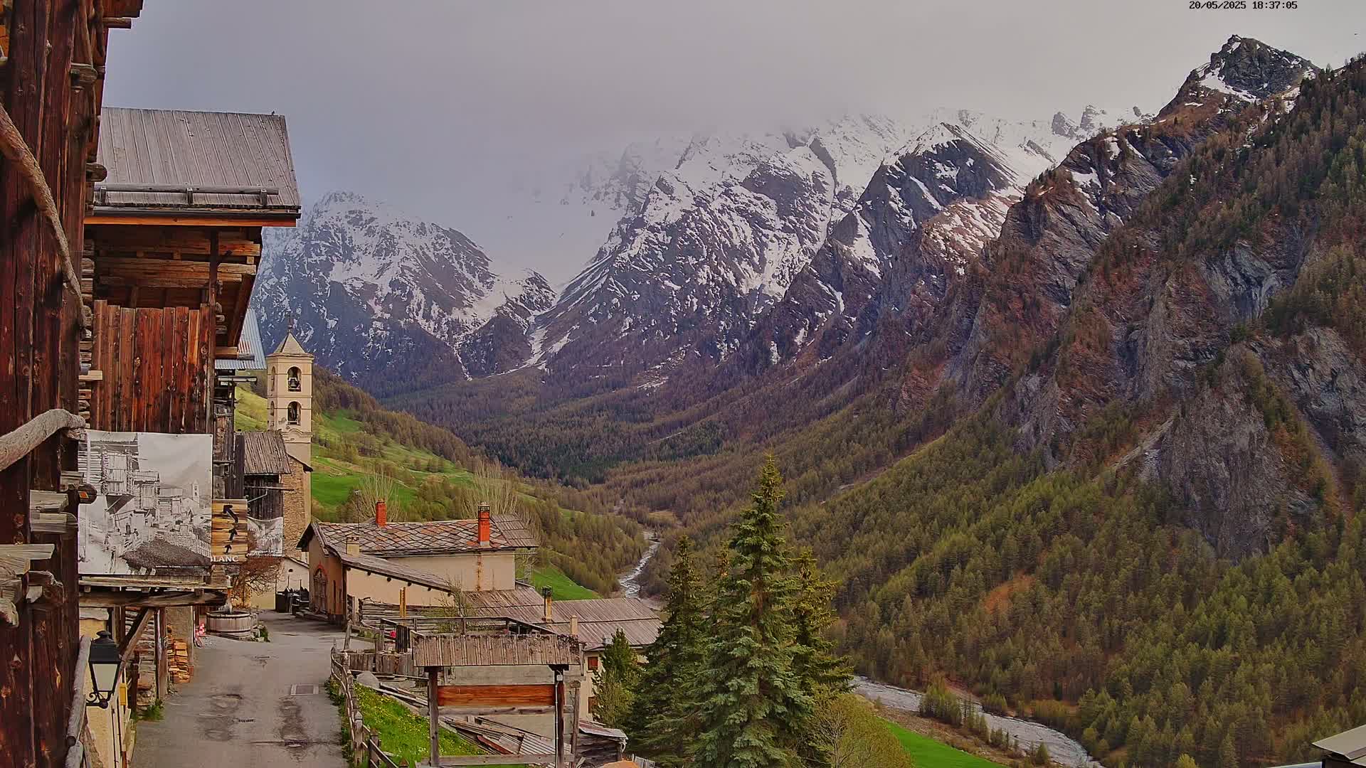 A view from a wooden building shows a small village nestled in a valley between snow-capped mountains on an overcast day.