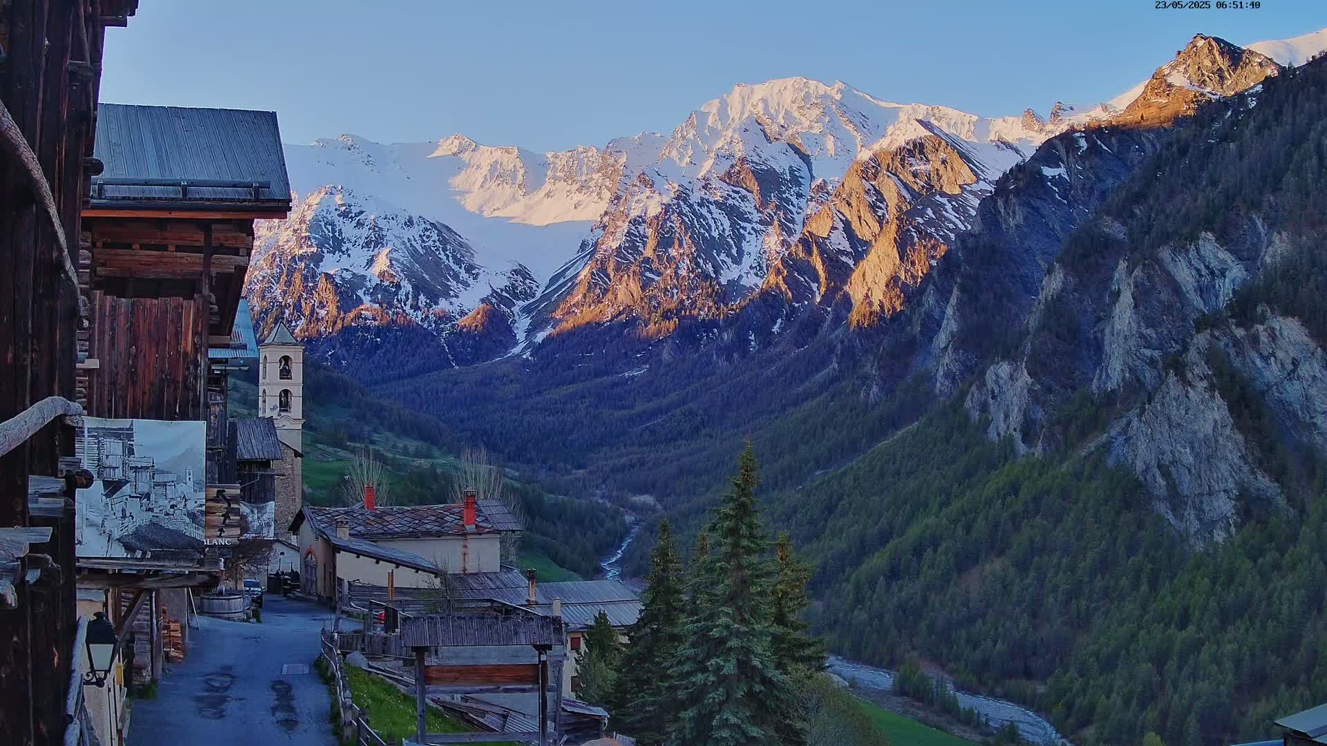 A quaint mountain village with old wooden buildings nestles in a valley, overlooked by a majestic, snow-capped mountain range bathed in the soft light of sunrise.