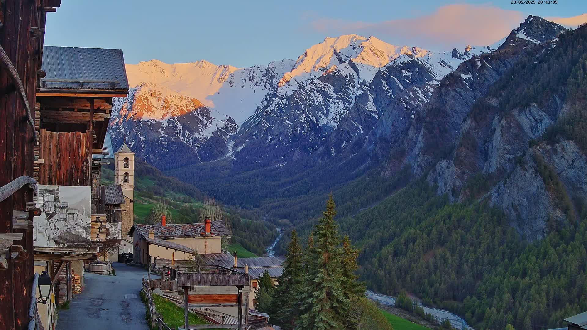 A quaint mountain village nestled in a valley at sunset, with snow-capped mountains rising in the background under a mostly clear sky.
