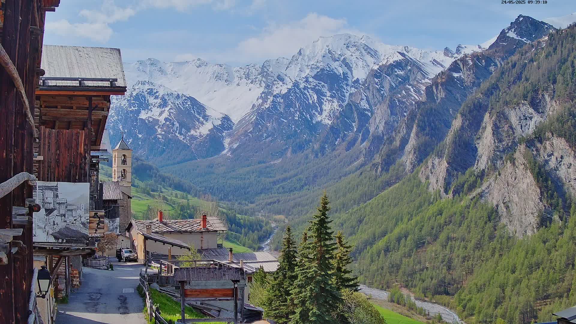 A quaint mountain village nestled in a valley, overlooked by snow-capped mountains under a partly cloudy sky.