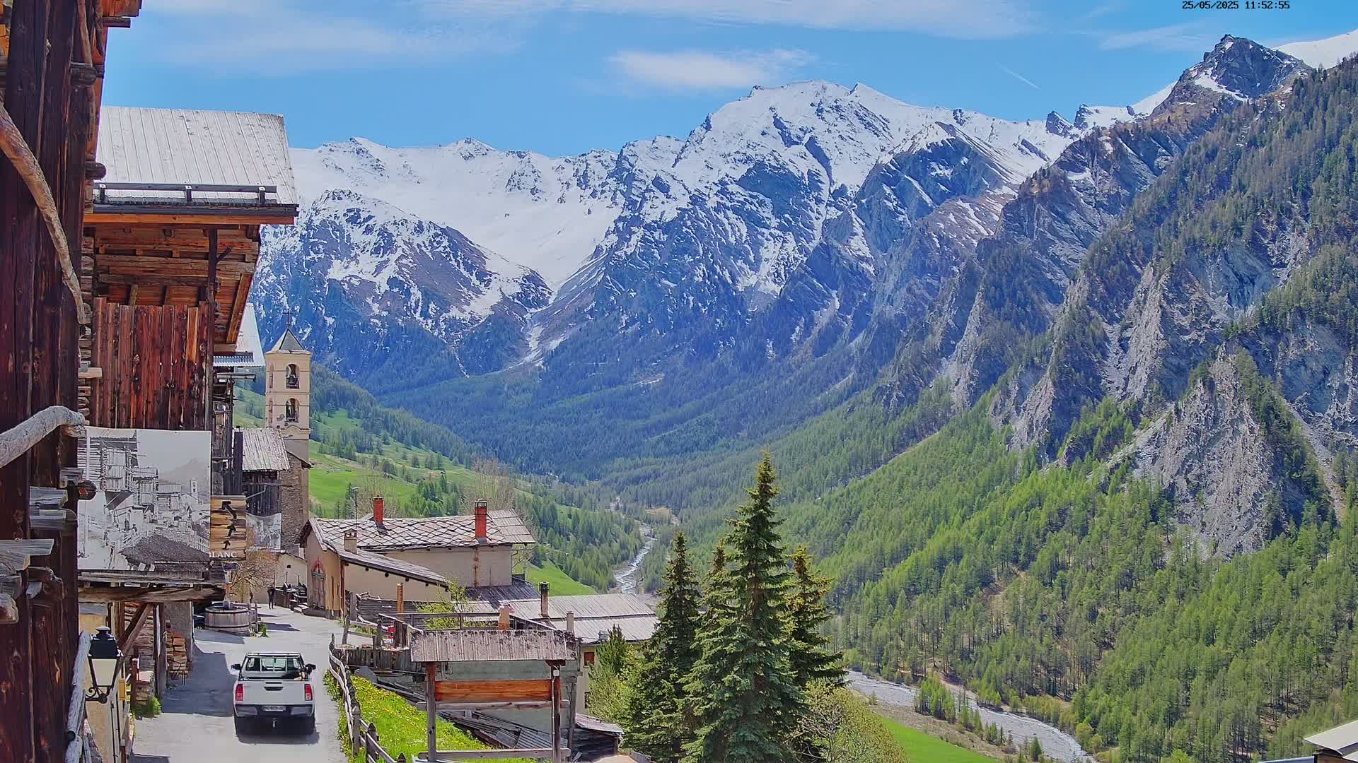 A sunny mountain village nestled in a valley, with snow-capped peaks in the background.