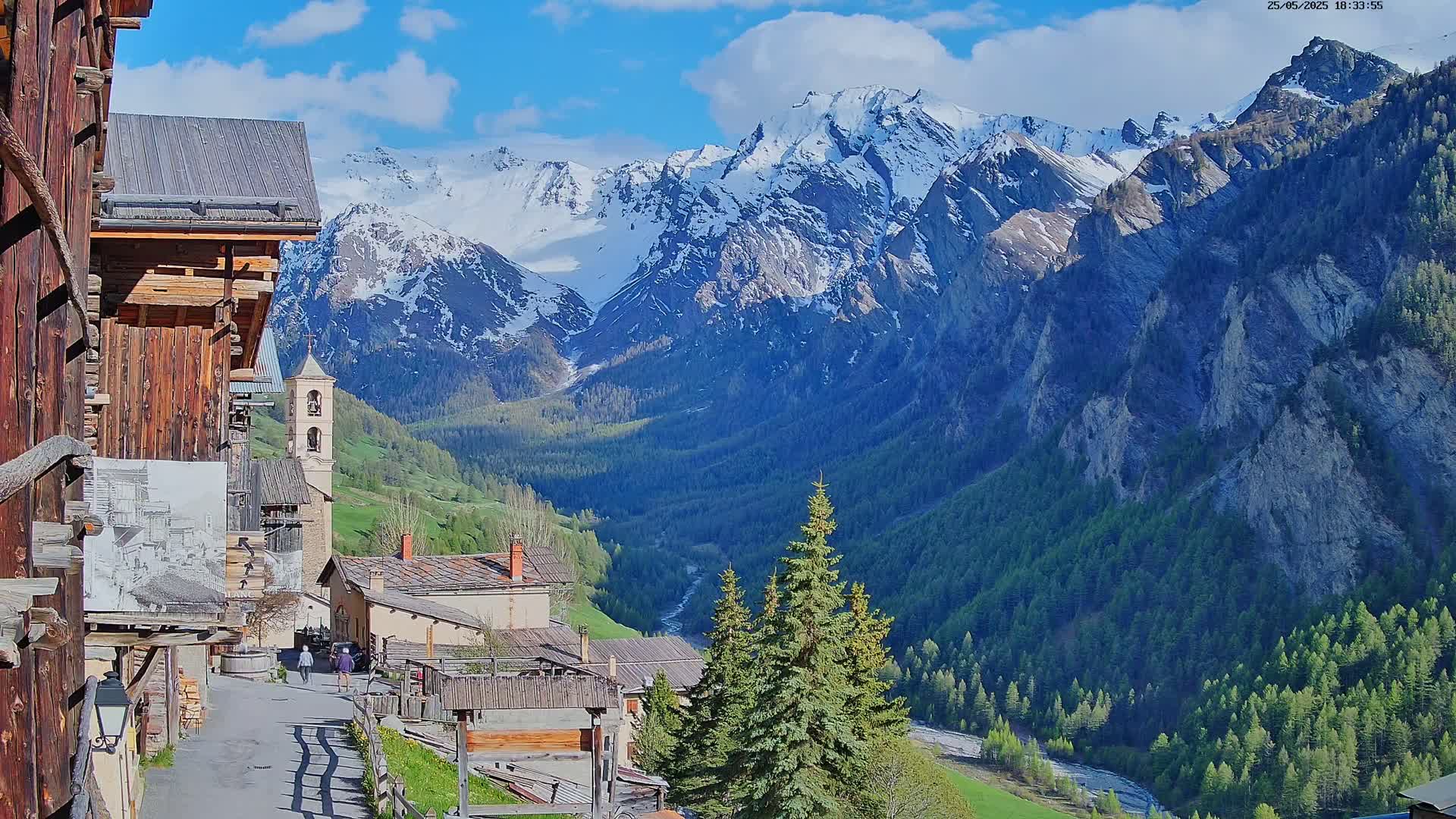 A quaint village nestled in a valley, with a church steeple visible, is overshadowed by a majestic, snow-capped mountain range under a partly cloudy sky.