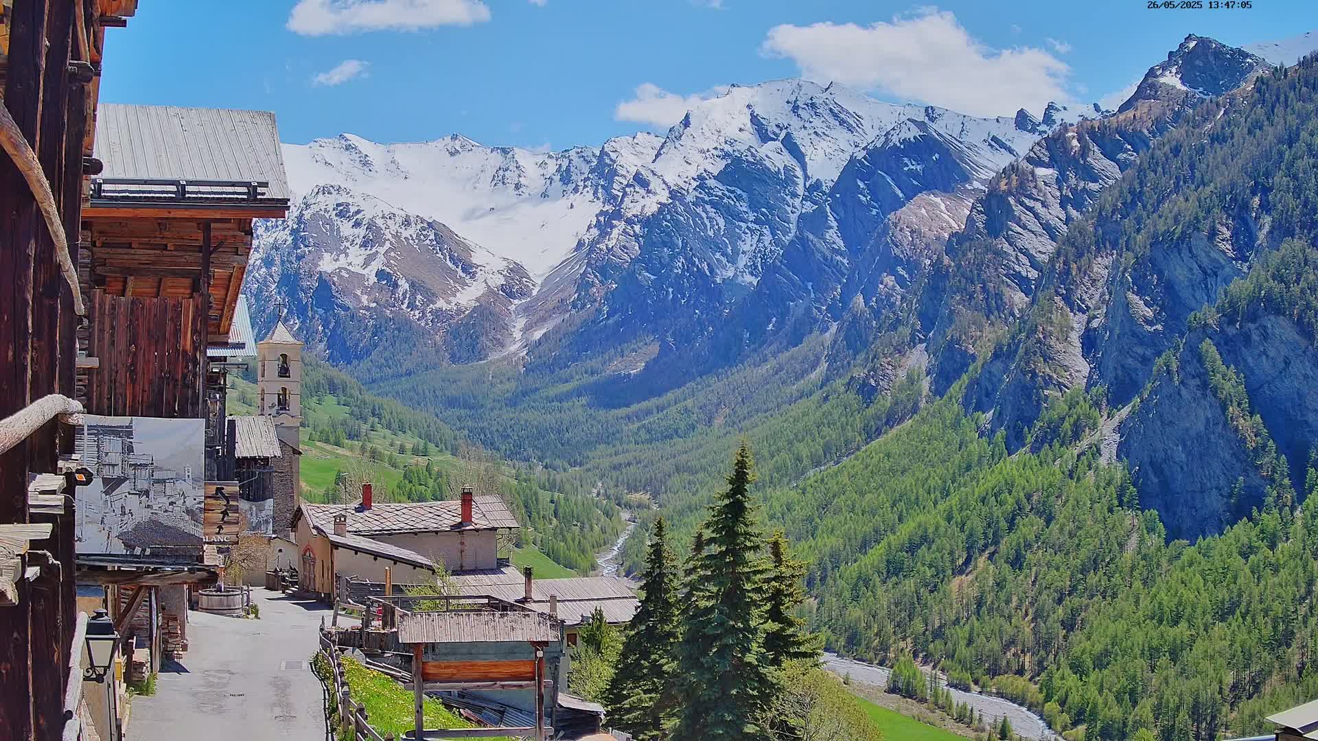 A quaint mountain village nestled in a valley, overlooked by snow-capped mountains under a partly sunny sky.