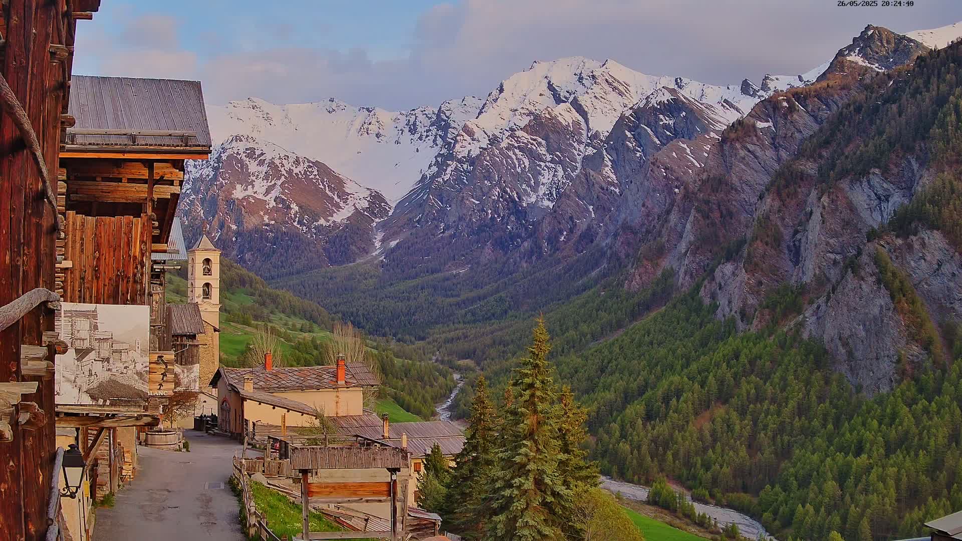 A quaint mountain village nestled in a valley, overlooked by snow-capped mountains under a partly cloudy sky.