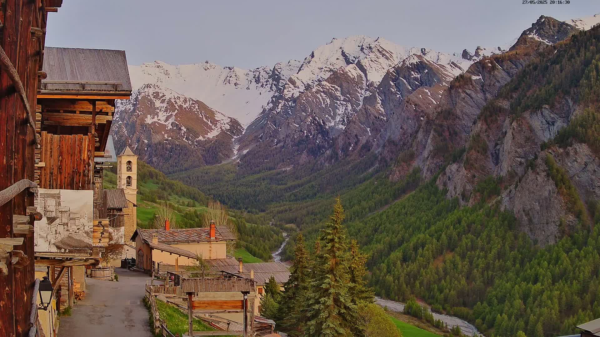 A quaint village nestled in a valley, surrounded by snow-capped mountains under a partly cloudy sky.