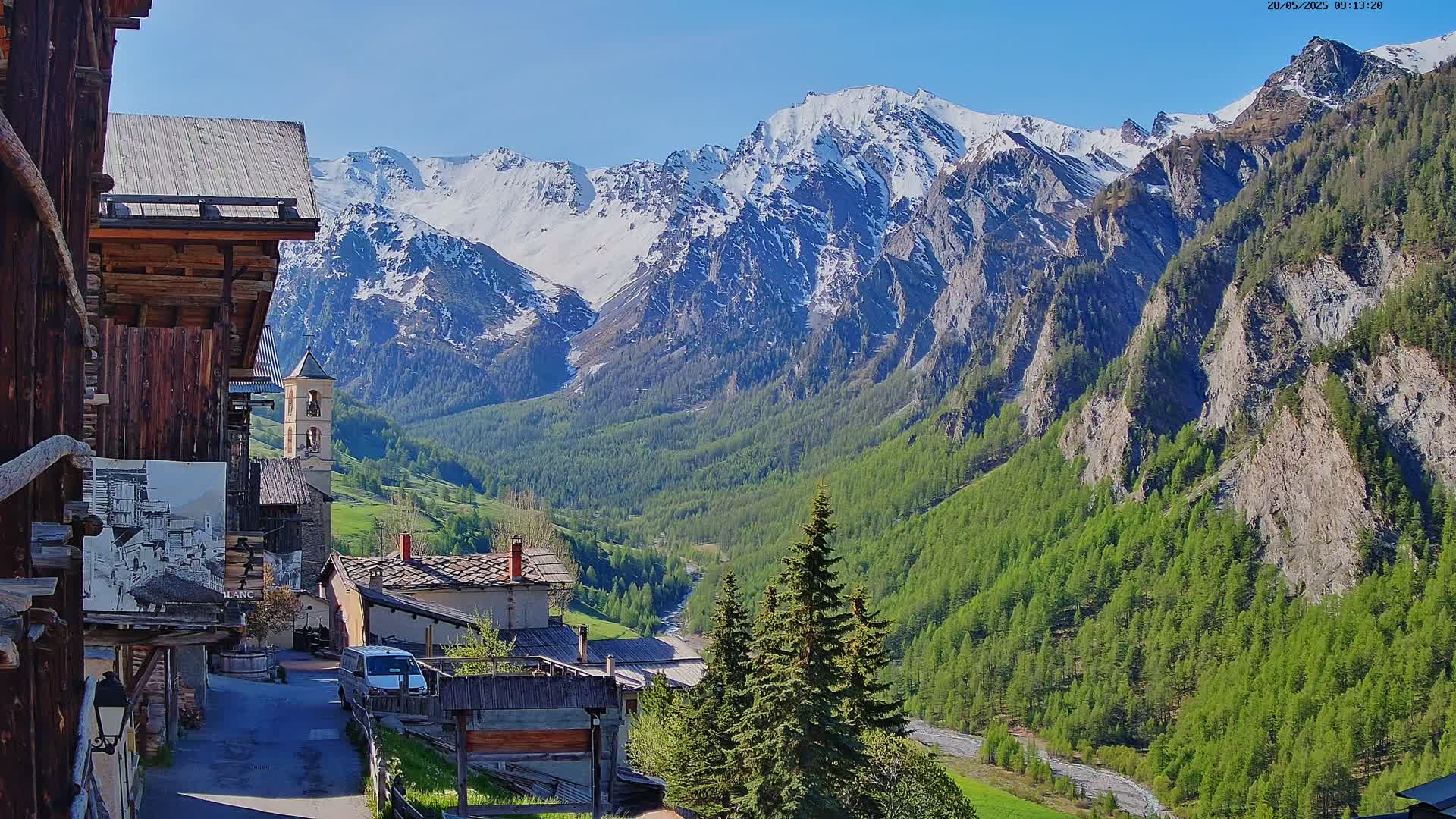 A sunny view of a village nestled in a valley between snow-capped mountains and lush green forests.