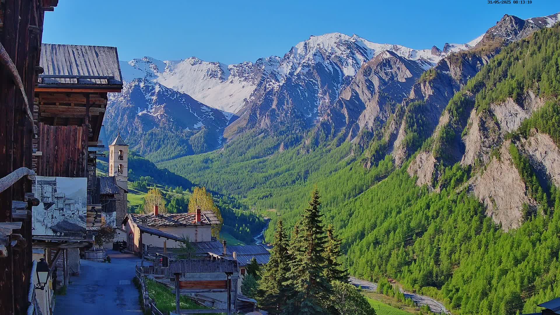 A quaint mountain village with old wooden buildings nestles in a valley beneath snow-capped mountains on a sunny day.