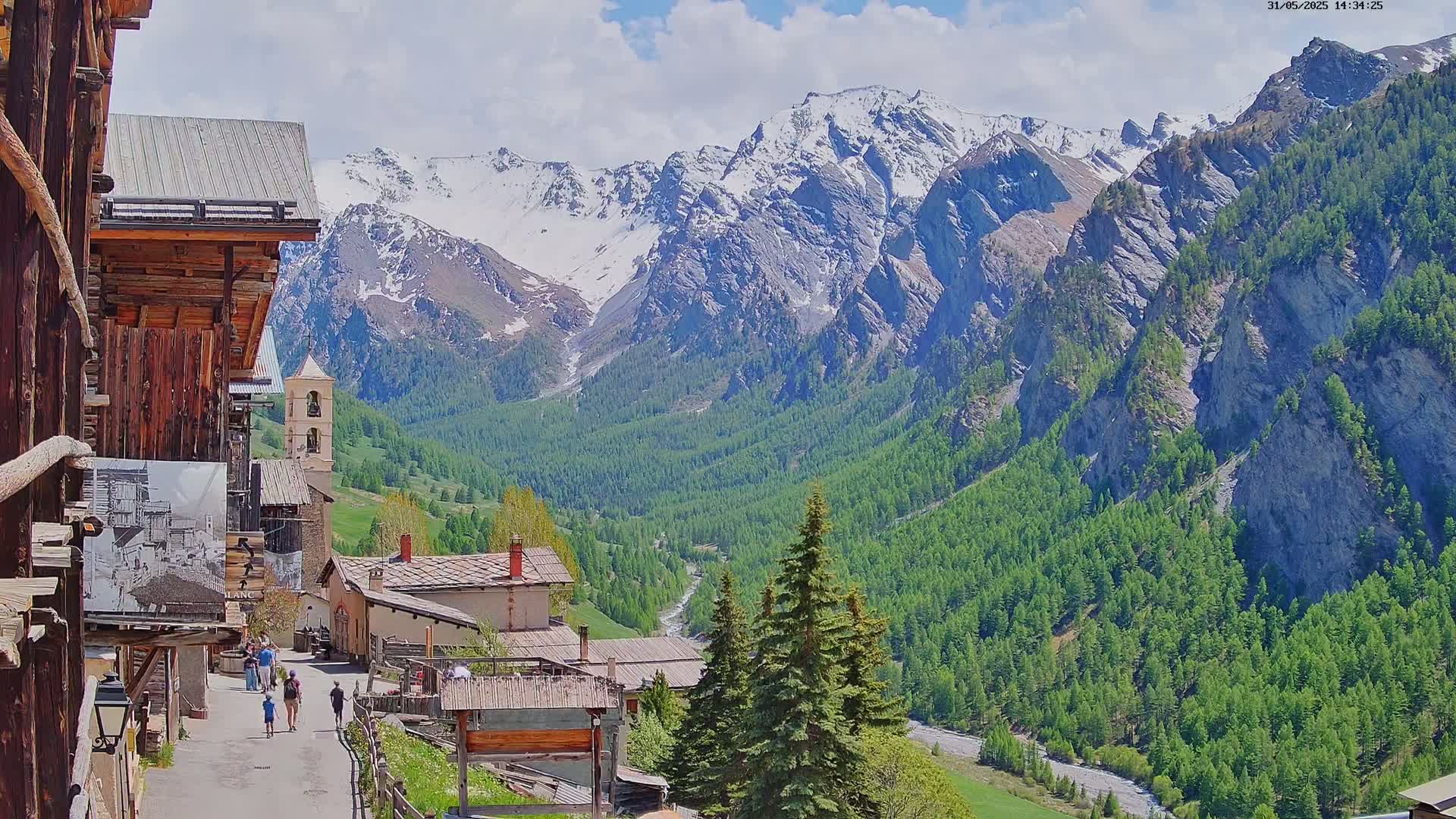 A picturesque mountain village with a bell tower, nestled in a valley surrounded by snow-capped mountains and lush green forests under a partly cloudy sky.
