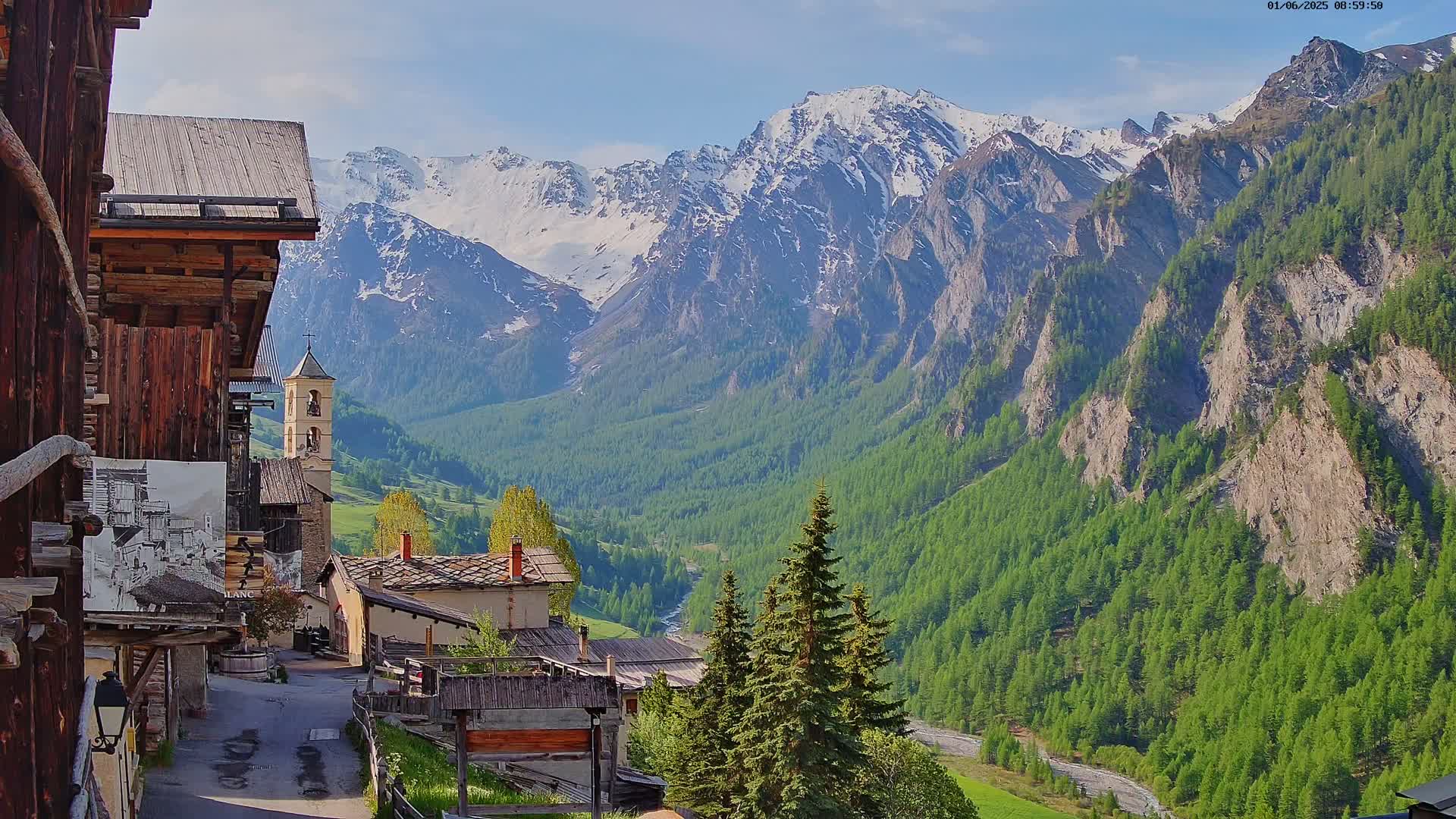 A quaint mountain village with wooden buildings nestles in a valley beneath snow-capped mountains on a sunny day.