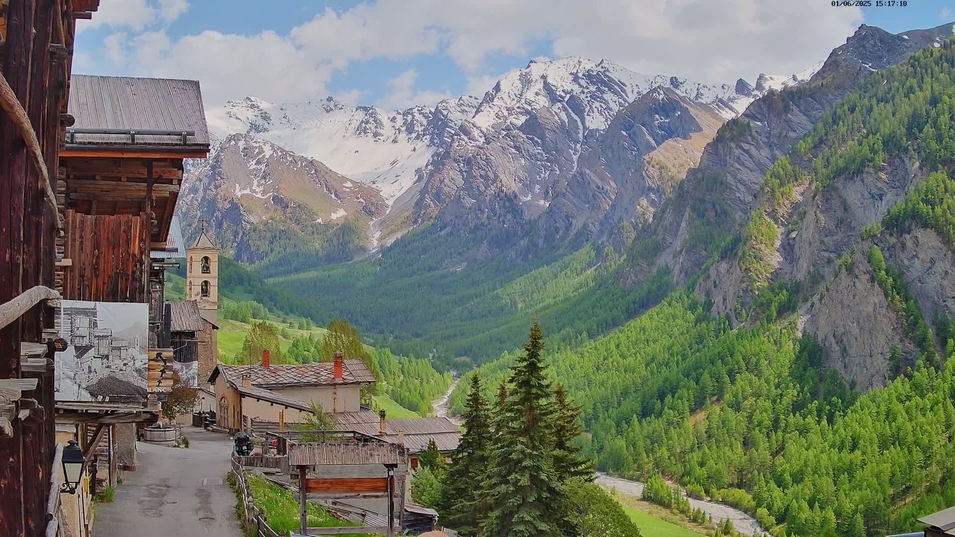 A picturesque mountain village nestled in a valley, with snow-capped mountains in the background under a partly cloudy sky.