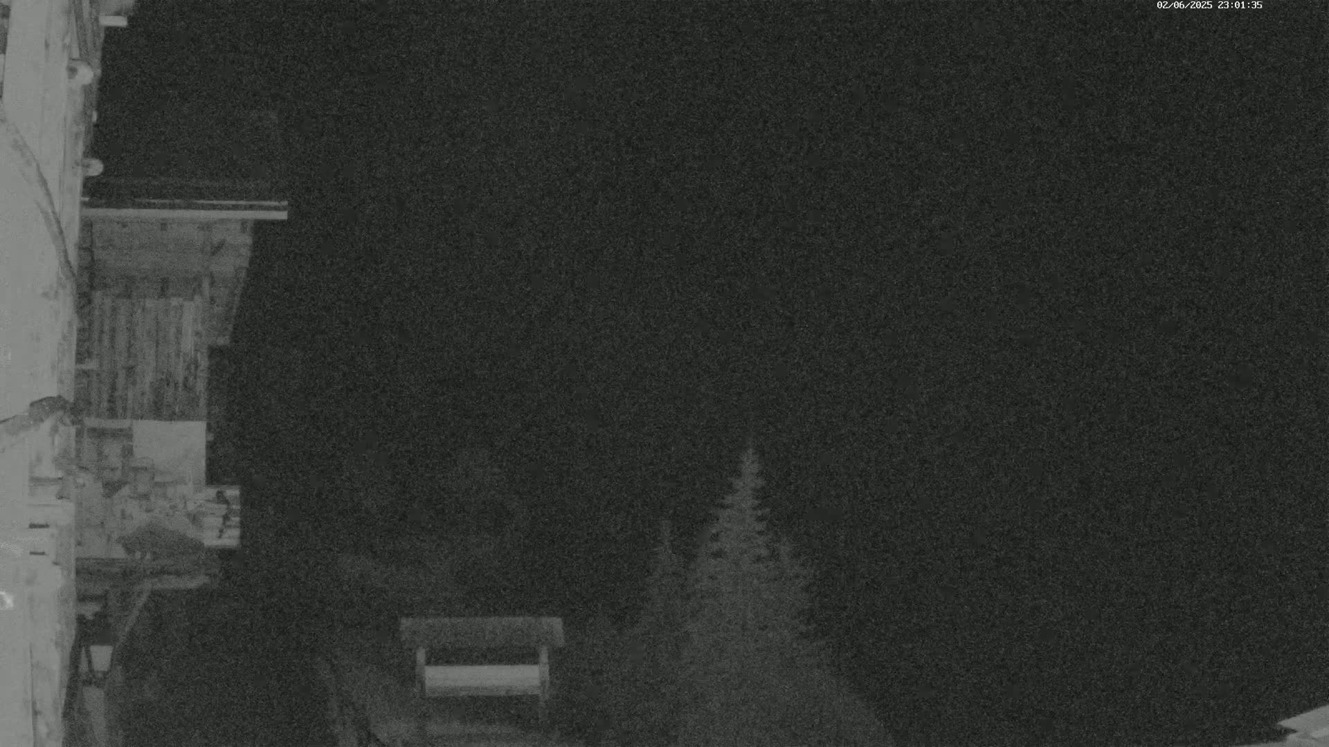 A nighttime, low-light image shows a portion of a building with a balcony overlooking a dark area with a few evergreen trees and a bench.
