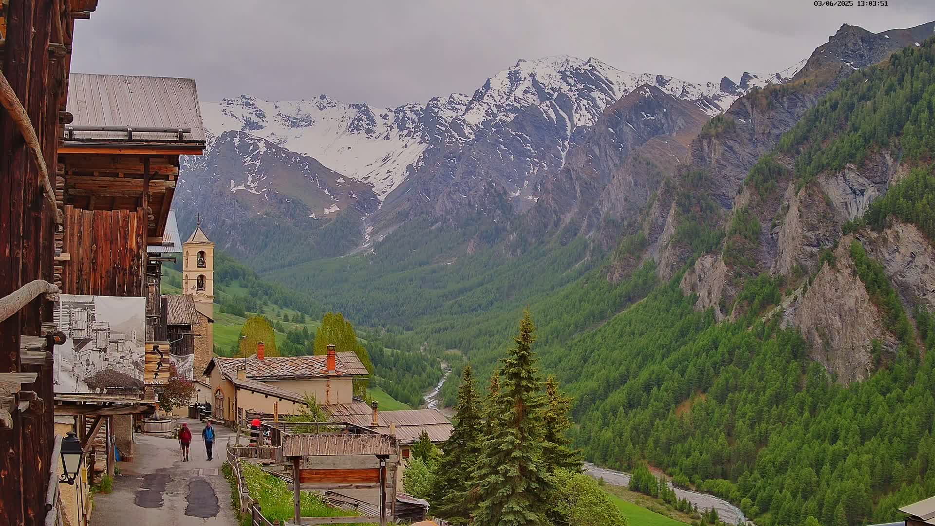 A quaint village nestled in a verdant valley, overlooked by snow-capped mountains under an overcast sky.