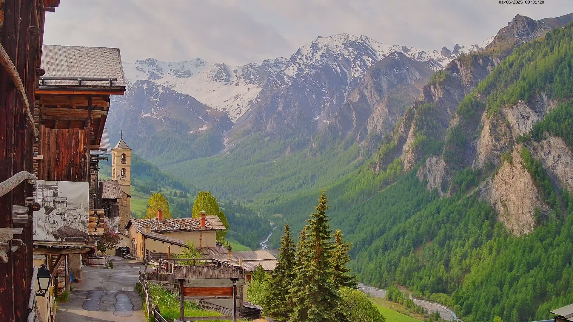 A quaint mountain village with a church steeple is nestled in a valley surrounded by snow-capped peaks and lush green forests under a partly cloudy sky.