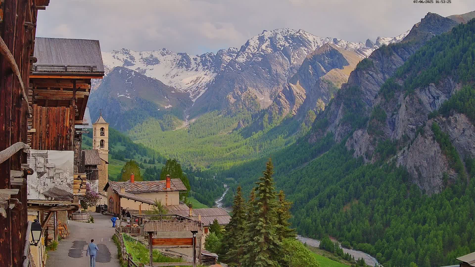 A quaint mountain village with wooden buildings nestles in a valley, overlooked by snow-capped mountains under a partly cloudy sky.